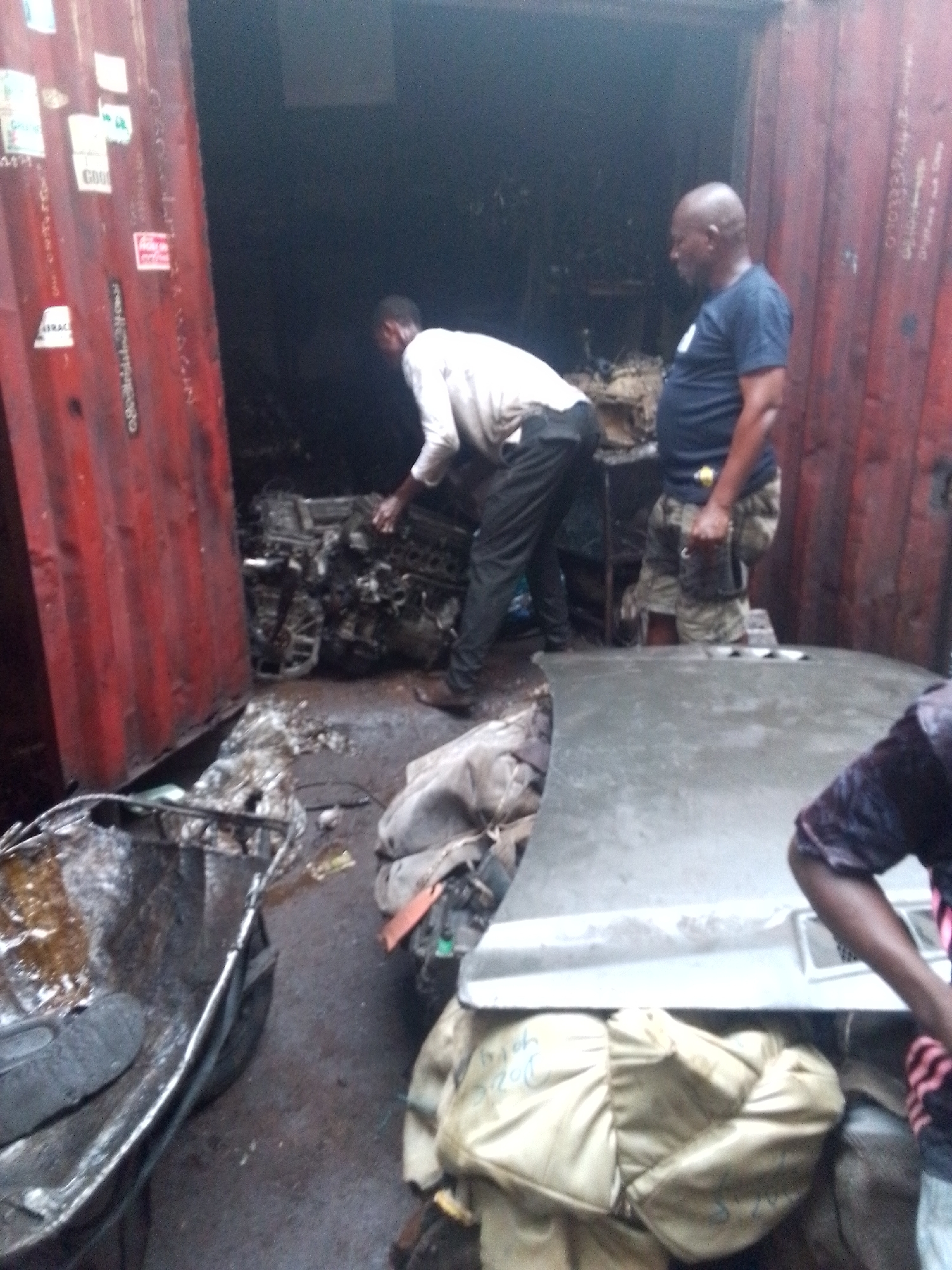 In a dimly lit shipping container in Onitsha, Nigeria, two men are inspecting a pile of car parts. The man on the left is wearing a white shirt and black pants, while the man on the right is wearing a navy blue t-shirt and camouflage pants. The container is filled with various car parts, including engines, hoods, and other salvaged components. There is also visible litter on the floor, making the environment appear cluttered and somewhat dirty. A third man is partially visible on the lower right corner of the image.