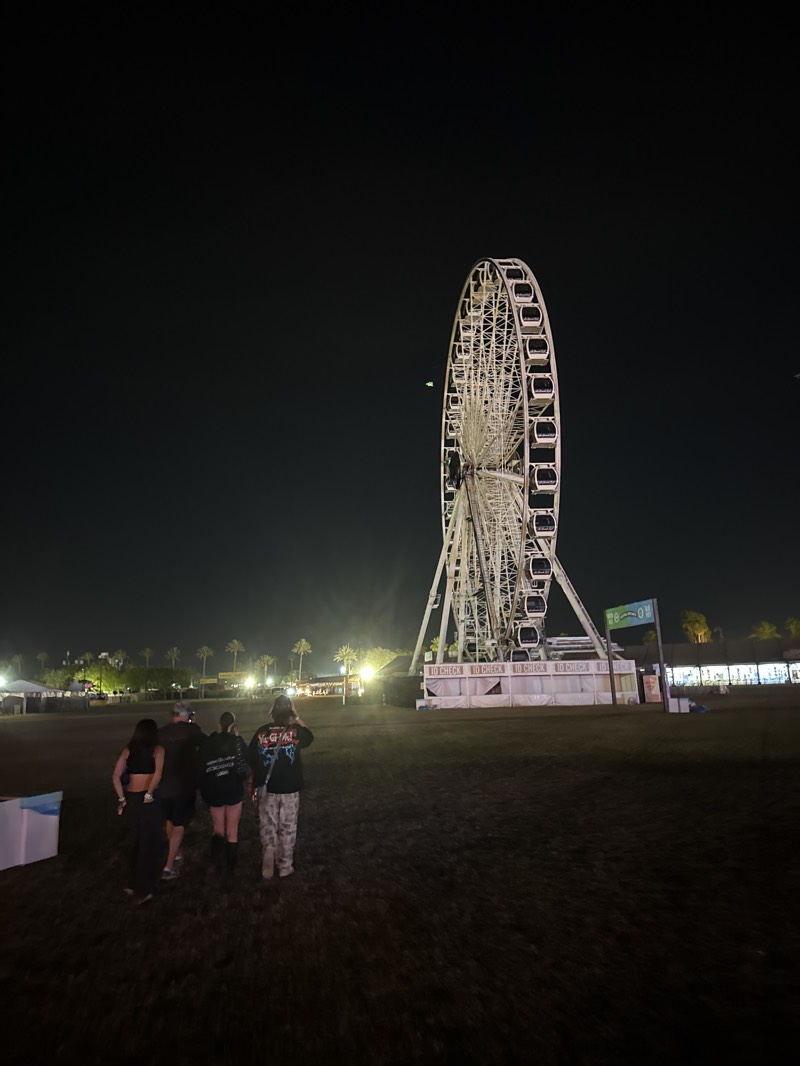 At night, a group of four people is walking on a grassy field in Indio, United States, towards a large, white Ferris wheel. The Ferris wheel is brightly lit and stands prominently in the background. In the distance, there are palm trees and buildings with lights. To the left, a blue object is present. The overall scene appears to be at an outdoor event or festival.