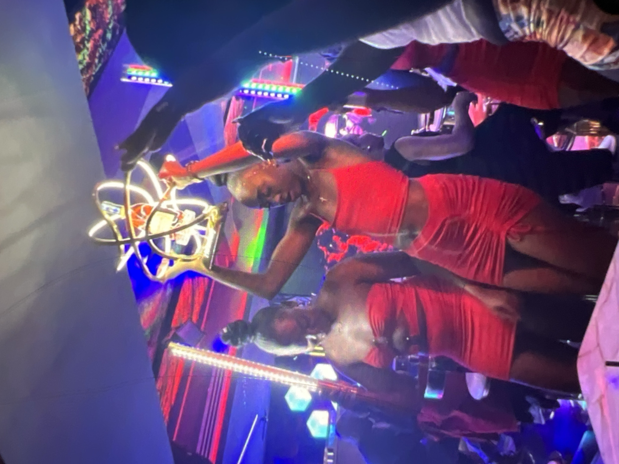 The image depicts a nightlife scene in Lagos, Nigeria, likely a club or party venue. Several individuals are visible, with the primary focus on two women in the foreground, both wearing red outfits. The woman on the left is wearing a strapless red dress, and the woman on the right is wearing a red crop top and a matching ruched skirt. The woman on the right is holding a golden, abstract sculpture above her head. The environment is dimly lit and illuminated by vibrant neon lights, characteristic of a lively urban entertainment setting. The background reveals more people and the general ambiance of a crowded social gathering.