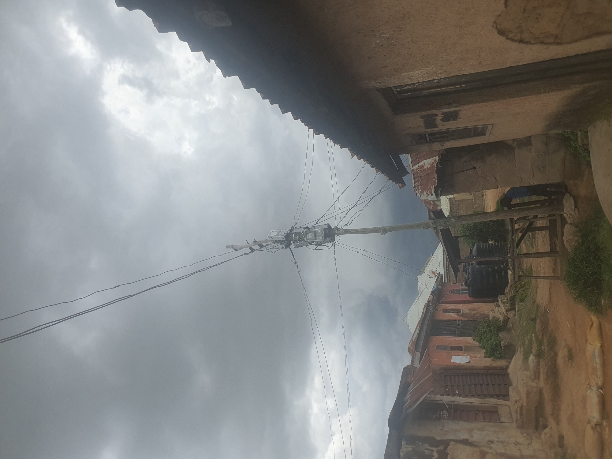 The image shows a street scene in a built-up area, likely a residential neighborhood, with a cloudy or overcast sky. The foreground features a dirt or unpaved road, with some grass and vegetation visible on the sides. Several buildings are present, constructed with materials that suggest a developing or informal settlement, possibly made of mud brick or concrete with corrugated metal roofing.
On the left, there's a structure with a weathered facade, a corrugated metal roof, and an open doorway. Adjacent to it is another building, painted an orange-red color, with barred windows and a corrugated metal roof. In the middle ground, a prominent utility pole stands tall, supporting electrical equipment including what appear to be transformers and junction boxes. Several electrical wires are strung across the scene, connecting to the pole and extending towards the buildings. Behind these structures, large black water tanks are visible.
To the right of the image, a building with a rough, textured wall, possibly mud, dominates the frame. It has a dark-colored door and a window. The roofline of this building is angled and appears to be made of corrugated material, extending over the side of the structure.
A person, wearing a blue garment, is partially visible near a wooden structure on the far right, appearing to interact with something on the ground or a low-lying object. The overall impression is of a functional, yet somewhat utilitarian, urban environment in Rukuba Road, Nigeria, under a subdued, possibly pre-rain, sky. The scene depicts a typical streetscape with essential infrastructure and residential dwellings.