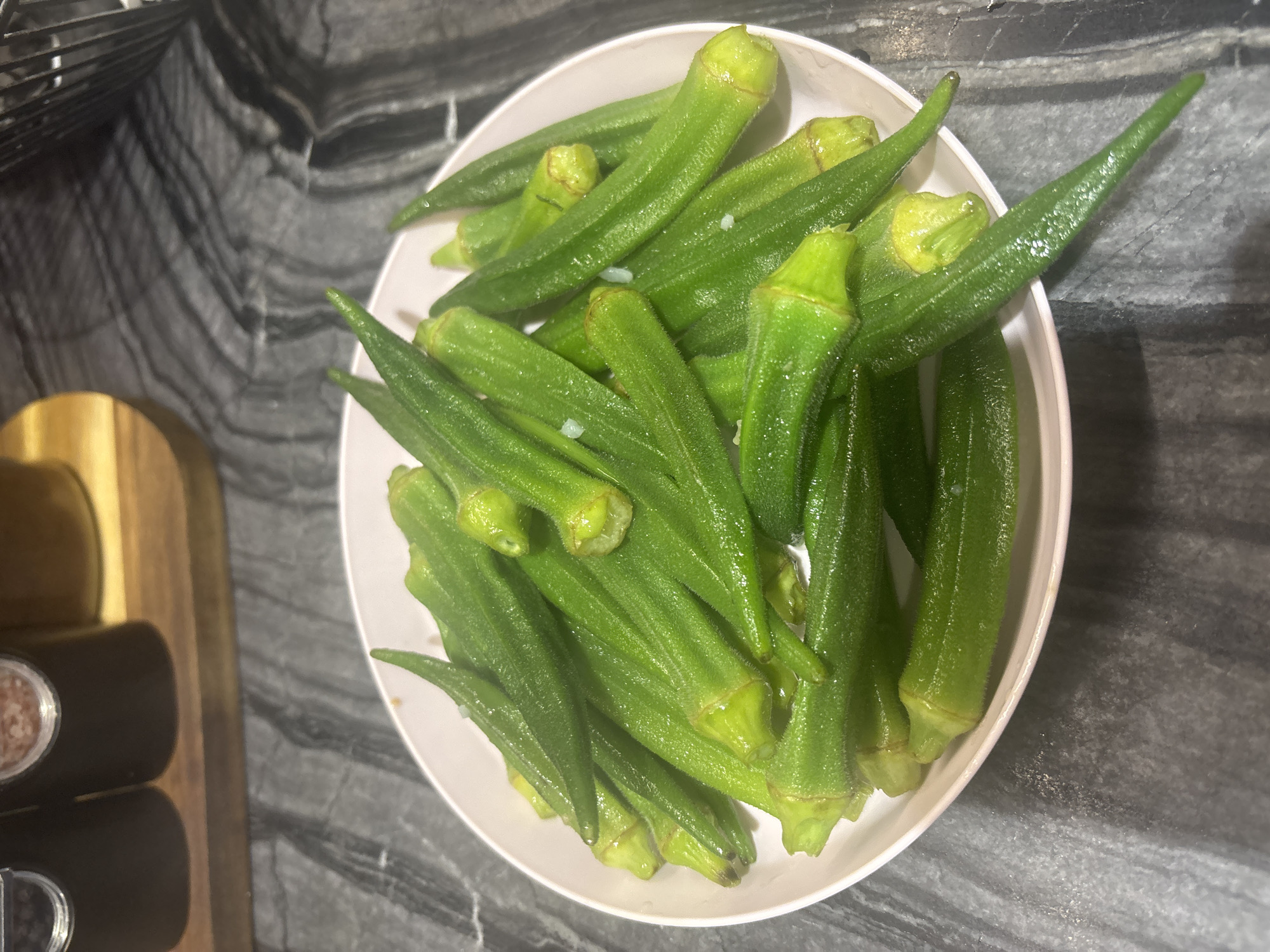 A white bowl filled with bright green okra sits on a dark, speckled countertop. The okra pods are whole and appear to be steamed or boiled, with a slight sheen from moisture. Some small white specks, possibly garlic or seasoning, are visible among the okra. In the background, out of focus, are two dark-colored spice grinders and a lighter-colored jar on a wooden cutting board. The overall scene is indoors, likely in a kitchen or dining area, with no indication of its specific location within Dubai, United Arab Emirates.