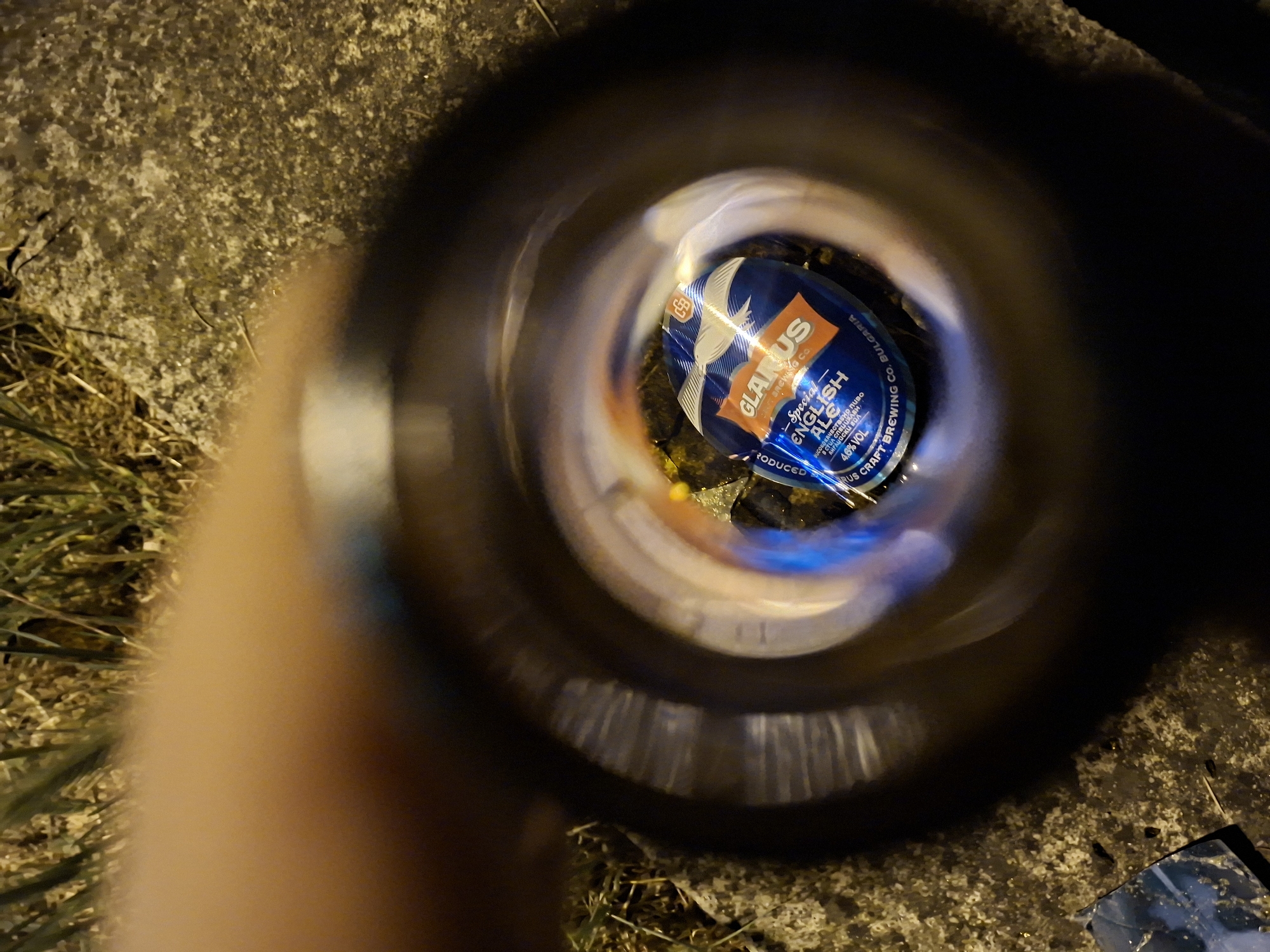 The image is a close-up, low-angle shot looking through the opening of a glass bottle. Inside the bottle, a blue and orange aluminum can is visible. The can has text that reads "GLARUS Special ENGLISH ALE" and "4.6% VOL." Below that, it says "CRAFT BREWING CO. BULGARIA." The can appears to be resting on a concrete surface, with some dry grass and dirt around it. The background is dark and slightly out of focus, with hints of green grass and speckled gray concrete. A blurry, flesh-toned object, possibly a finger or hand, occupies the top left portion of the frame, obscuring part of the scene. The lighting suggests it is nighttime or a dimly lit environment. The scene is likely located outdoors, possibly in a public space in Varna, Bulgaria, given the mention of "BULGARIA" on the can.
