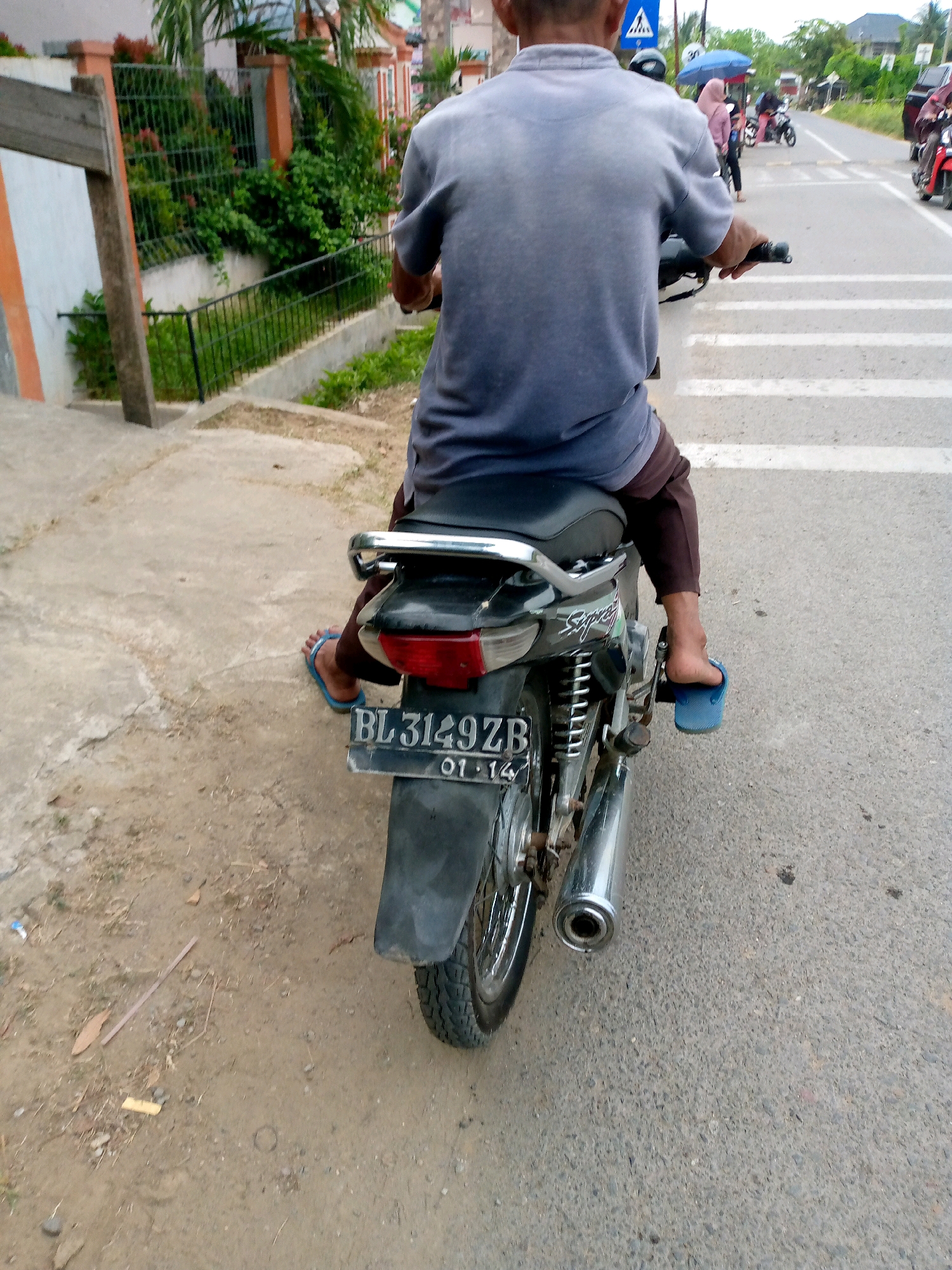 The image shows a person riding a motorcycle on a street in Mesjid, Indonesia. The rider is wearing a gray t-shirt and brown pants, and blue flip-flops. The motorcycle is black with a silver exhaust pipe and a license plate that reads "BL 3149 ZB 01-14". In the background, there are buildings, trees, and other motorcycles. The street appears to be paved, and there are white lines indicating a crosswalk. The weather is overcast.