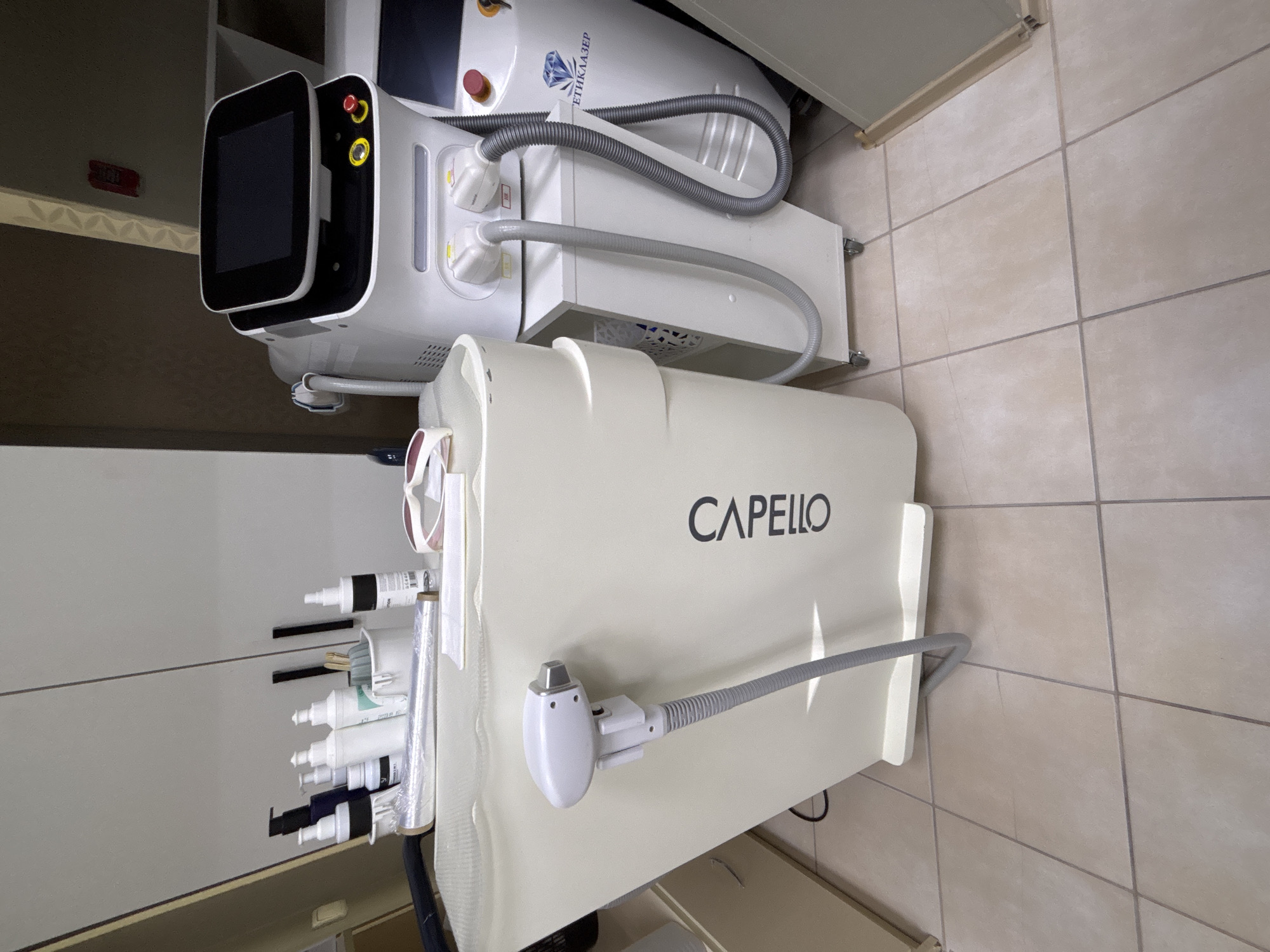 The image shows two medical or cosmetic treatment devices situated indoors, likely in a clinic or treatment room. One device features a black touchscreen display and several knobs, while the other is labeled "CAPELLO" in bold, sans-serif lettering. Various tubes, bottles, and accessories are attached to or located near the devices. The floor is tiled, and the lighting appears to be artificial. While the prompt indicates the location is Vsevolozhsk, Russia, there is no inherent indication of geographical location in the image itself.