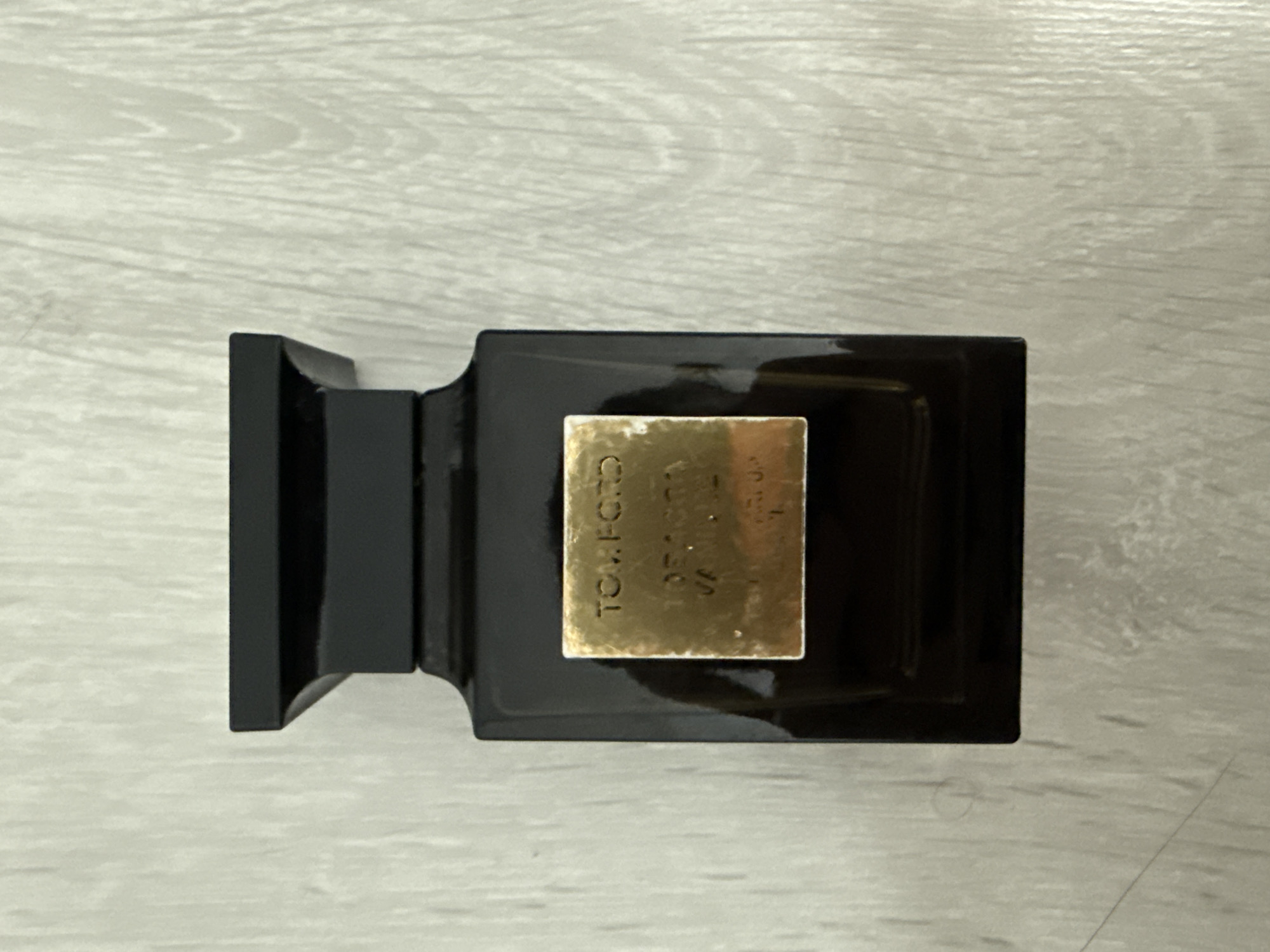 A black rectangular Tom Ford perfume bottle with a gold label is centered on a light-colored wooden surface. The label is partially obscured by a reflection, but the words "TOM FORD" are clearly visible at the top. Below that, some text is visible, including "TOBACCO" and "VANILLA". The wooden surface appears to be a table or floor with a light wood grain pattern. There are no people or other objects in the image. The location of Burgas, Bulgaria is not discernible from the image.