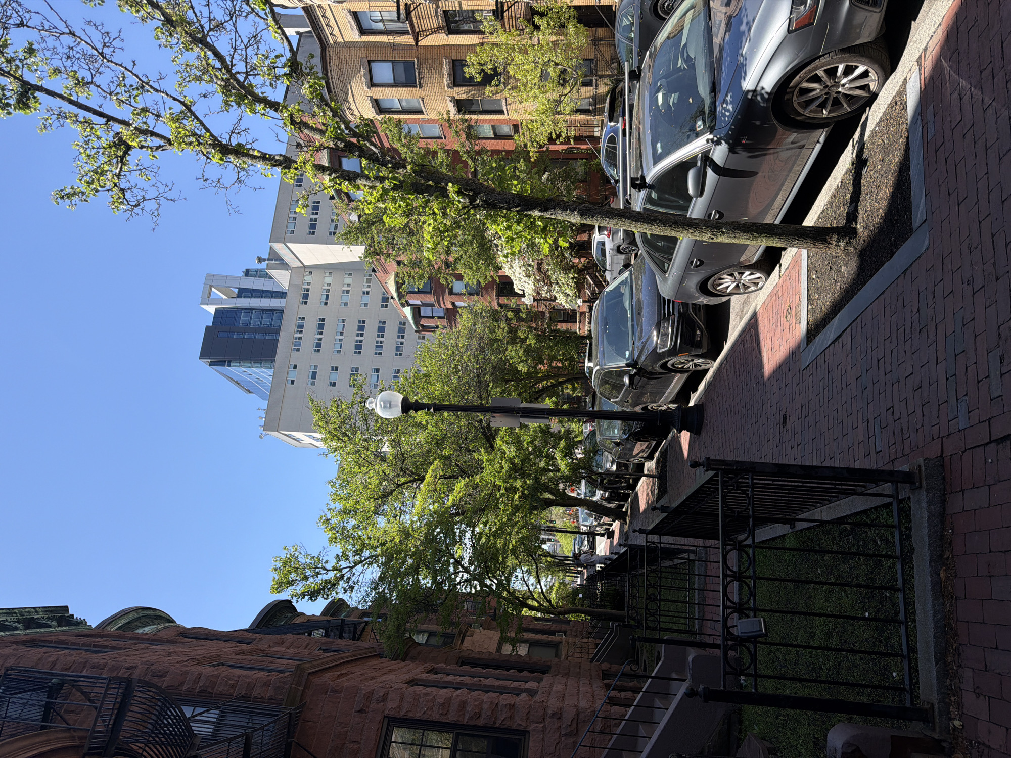 The image depicts a street scene in Boston, USA. A row of parked silver and gray cars lines the right side of the street along a red brick sidewalk. On the left, brownstone buildings with visible fire escapes and black wrought iron fences are present. Tall trees with green leaves line the street, casting shadows. A black lamppost stands among the trees. In the background, taller buildings and a clear blue sky are visible.