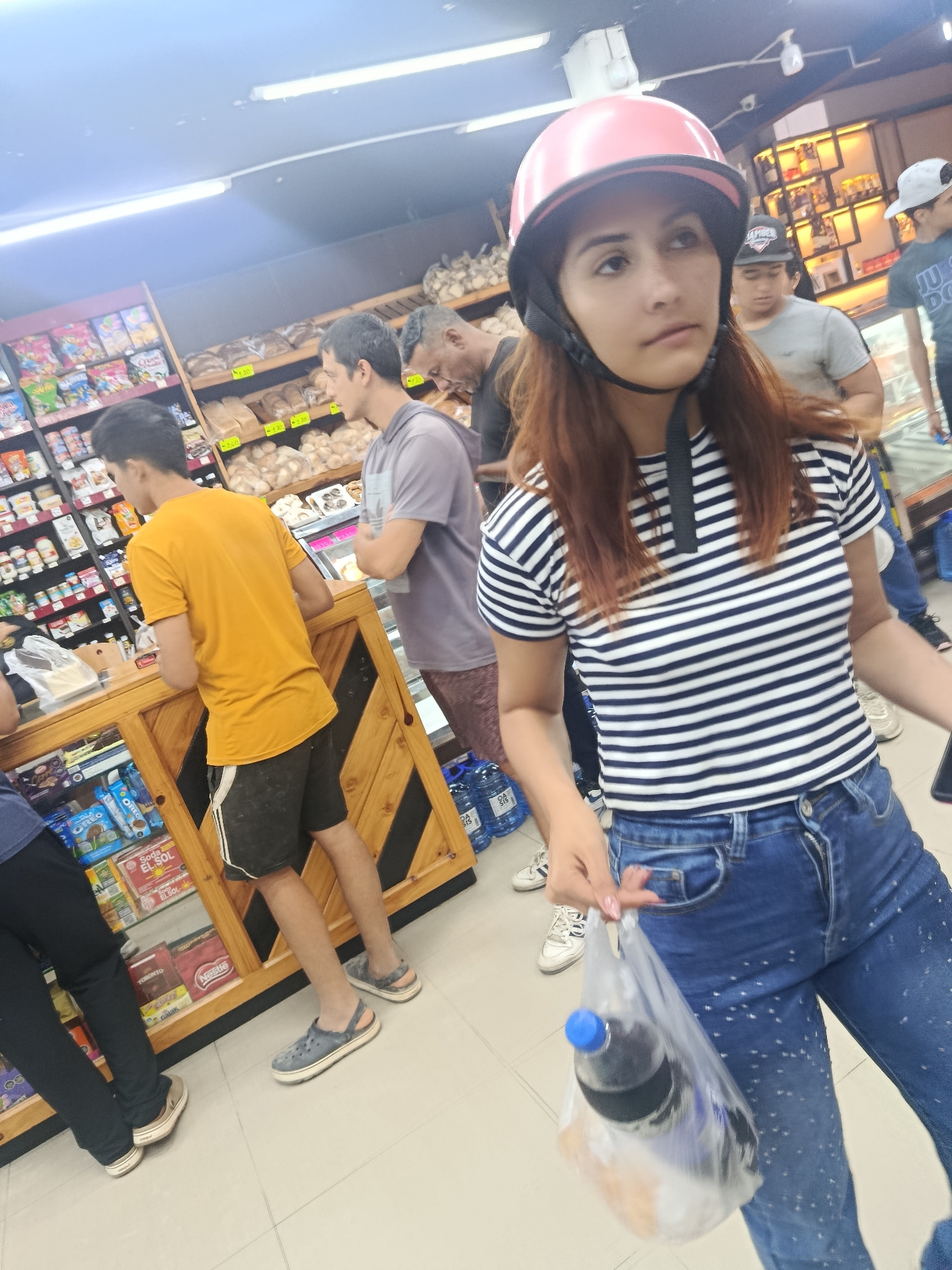A woman with long reddish-brown hair wearing a pink helmet, a striped shirt, and blue jeans holds a plastic bag containing a bottle of soda in a store. She stands in a brightly lit interior space with a tiled floor, possibly a grocery or convenience store, based on the shelving stocked with various goods visible in the background. Other people are present, including some waiting by a counter or display case containing snacks, and others standing further back towards the shelves. The location is Altagracia, Venezuela.