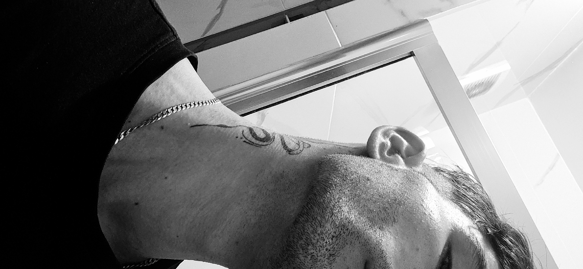 This black and white, high-contrast image presents a close-up, low-angle perspective of a person's upper body and head, likely taken in a modern indoor setting, possibly a bathroom in Plovdiv, Bulgaria.
The main subject is a person, appearing to be male, whose profile is captured with their head tilted downwards and to the side. They are wearing a dark, possibly black, t-shirt. A prominent silver chain necklace is visible around their neck. On the side of the neck and extending towards the shoulder, there is a tattoo with abstract or stylized designs. Short stubble is visible on the person's cheek and jawline, and dark hair is seen near the forehead.
The background features light-colored, possibly white, rectangular tiles and a reflective surface, likely glass, encased within a light-colored or metallic frame. These elements suggest a shower enclosure or a large mirror. The bright lighting creates strong contrasts and highlights, especially on the person's skin and the reflective surfaces. No specific activity is taking place, and the person appears to be stationary. No emotions are discernible from the visible parts of the face, and there is no visible text in the image.