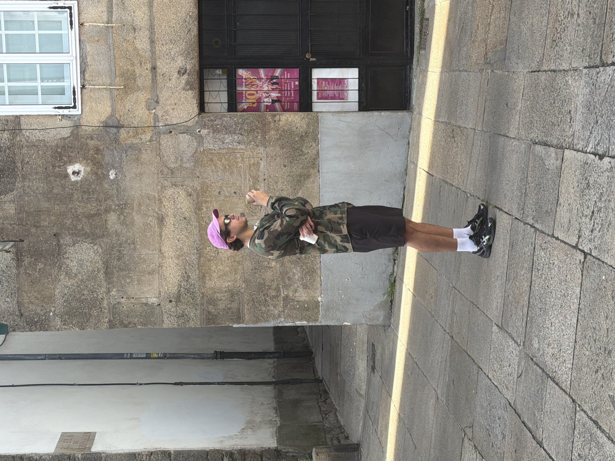 The image depicts a young person standing on a cobblestone street. The person is wearing a camouflage jacket, black shorts, white socks, and black sneakers. They are also wearing sunglasses and a pink baseball cap. The person is holding a white object, possibly a phone or a cup, in their right hand.
The background features a stone building with a large black door and a window with posters. To the left of the door, there is a white window with multiple panes. The street is paved with large, irregular granite stones. Sunlight casts long shadows across the pavement, suggesting it is either morning or late afternoon. The scene appears to be in a European city, consistent with the given location context of Santiago De Compostela, Spain. The overall mood of the image is casual and relaxed.