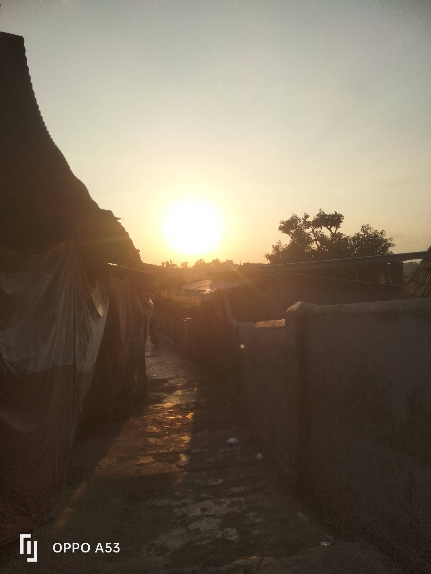 The image depicts a street scene at sunset in Badung, Indonesia. The sun, a bright orb, hangs low in the sky, casting a warm glow and long shadows. The street is narrow, flanked by buildings on either side. On the left, a structure covered with what appears to be tarp or fabric is visible. On the right, a wall made of concrete or brick runs along the street. The ground is paved, with some areas appearing wet or reflective, suggesting recent rain or puddles. In the distance, trees are silhouetted against the sky, and the roofs of other buildings are visible. The overall atmosphere is peaceful and serene, with the setting sun creating a dramatic and colorful backdrop. The image also includes the branding "OPPO A53" in the bottom left corner.