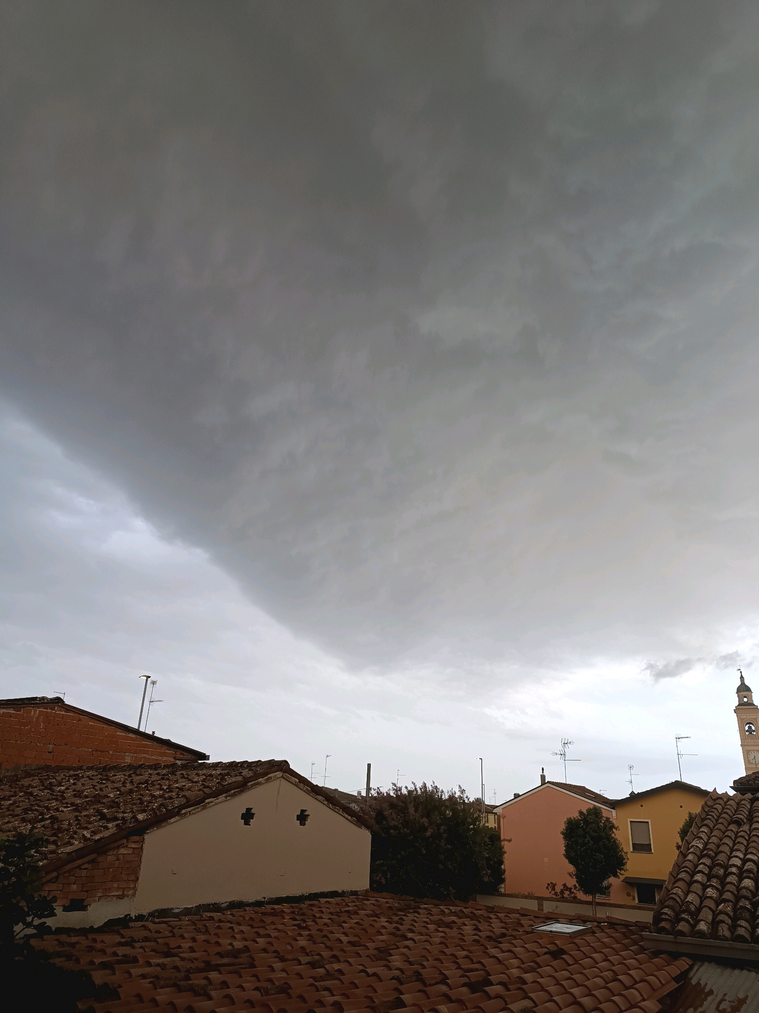 The image captures a cloudy sky over a cityscape. In the foreground, terracotta tiled roofs are visible, suggesting a European setting. Behind the roofs, several buildings of varying colors, including brick red, cream, pink, and yellow, can be seen. The sky is dominated by dark, heavy clouds, indicating an impending storm or overcast weather. A distant clock tower with a bell can be spotted to the right. The scene appears to be a view from a rooftop in the city of Viadana, Italy, characterized by its typical Italian architecture and rooftops. No people are present in the frame.