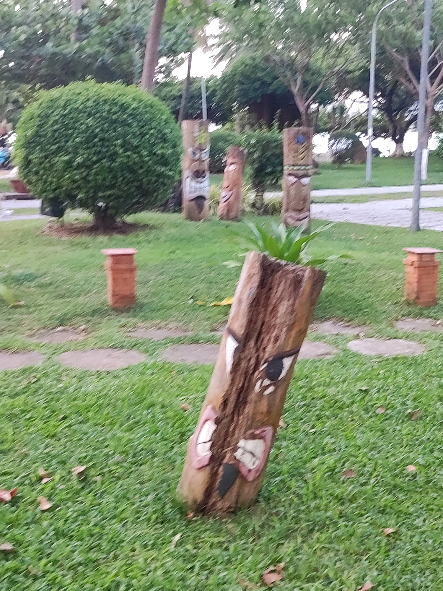 The image depicts a grassy park area with several carved wooden posts resembling tiki statues. The statues are decorated with painted faces, featuring exaggerated eyes and mouths. One carved log in the foreground is tilted and has a small plant growing out of its top. In the background, a neatly trimmed, spherical bush stands out against a backdrop of trees and a streetlamp. Scattered throughout the grass are a few fallen leaves. Two small, cylindrical brick structures are visible, one on the left and one on the right side of the frame. The scene appears to be an outdoor public space, possibly a park or garden. There is no visible text in the image. The lighting suggests it is daytime, and the overall atmosphere is calm and serene. The location is identified as Nha Trang, Vietnam.
