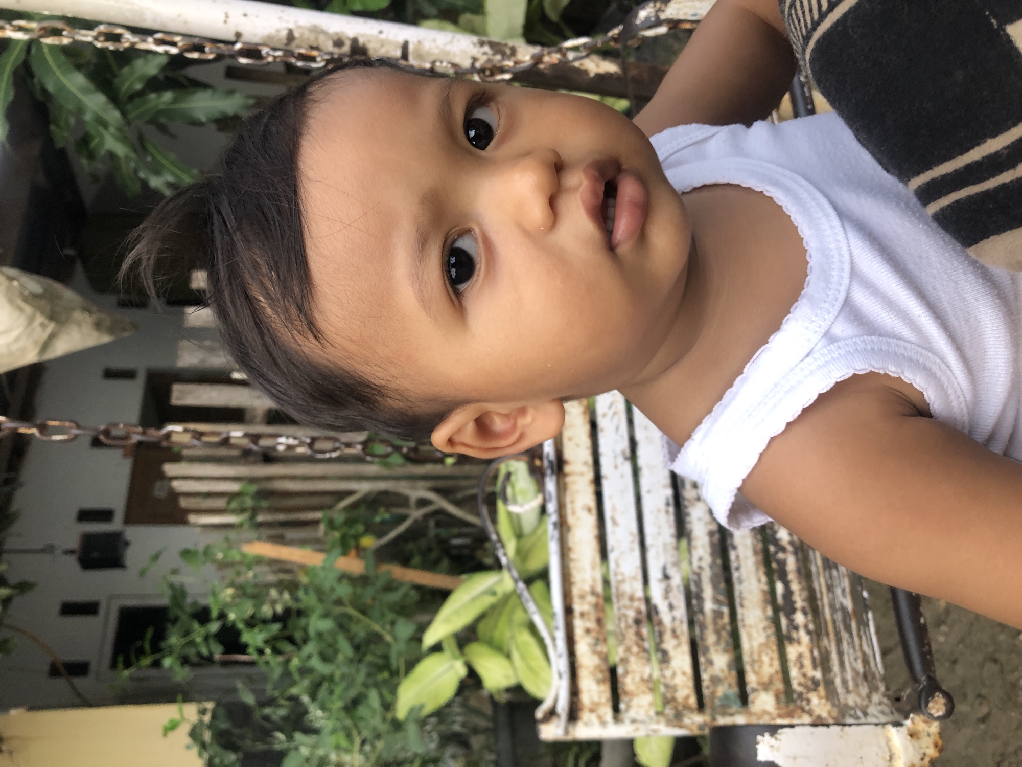 A close-up, slightly angled shot of an infant with dark hair, wearing a white tank top, sitting on an aged, metal swing-like chair. The swing is set against a background of outdoor foliage and part of a building. The location is unknown. The infant is facing the camera, with their gaze directed at the viewer. The setting appears to be outdoors during daylight.