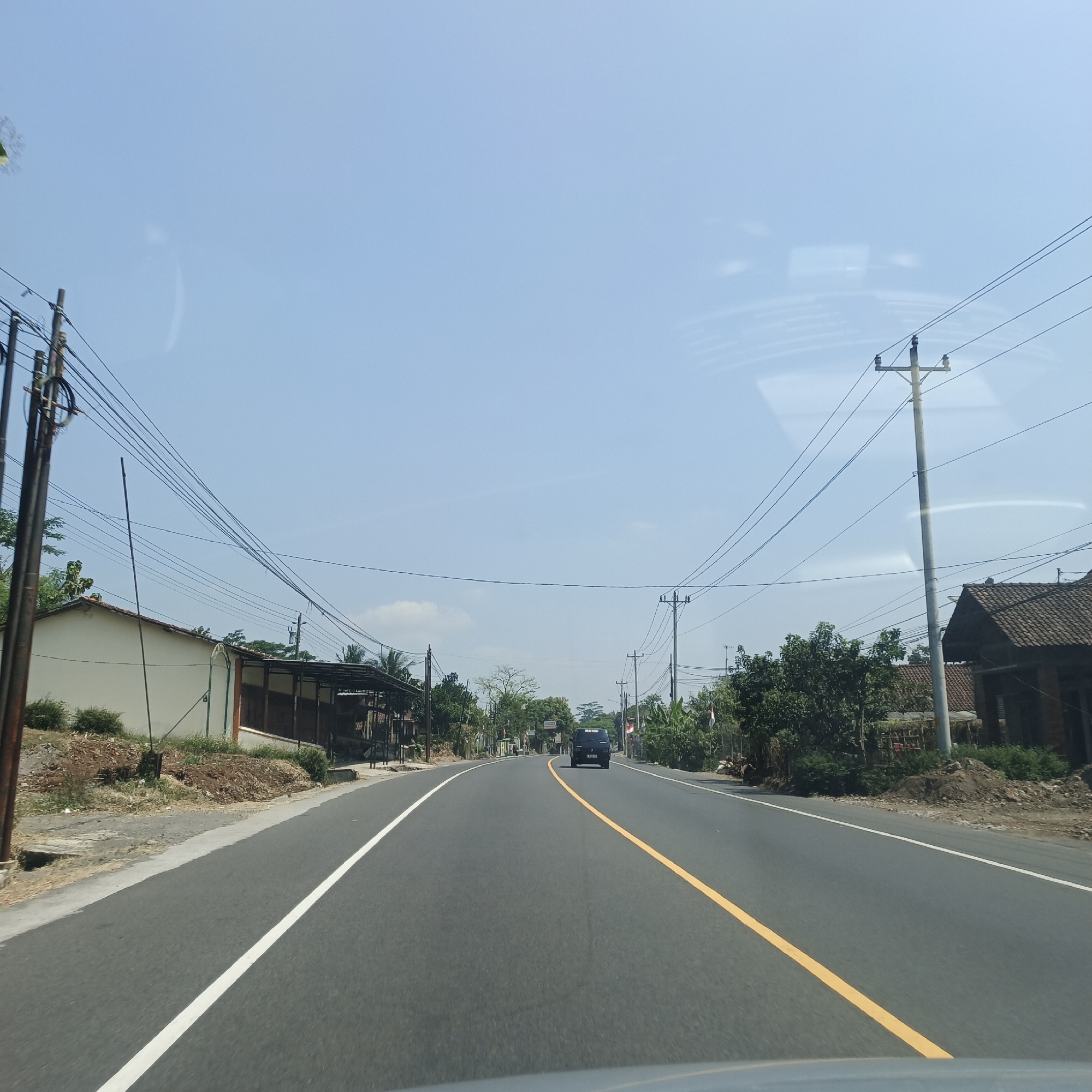 The image depicts a paved, two-lane road in Soropadan, Indonesia, under a clear blue sky. The road features a solid white line on the left and a broken yellow line on the right, dividing the lanes. A dark-colored SUV is visible driving away from the camera in the distance. The road is bordered by a dirt shoulder on the left, with a small building and sparse vegetation. On the right, there are more trees, some housing with tiled roofs, and utility poles with numerous electrical wires crisscrossing overhead. The environment appears to be rural or semi-rural, with evidence of infrastructure and residential buildings lining the roadway. The lighting suggests it is daytime, with the sun casting shadows from the utility poles and buildings.
