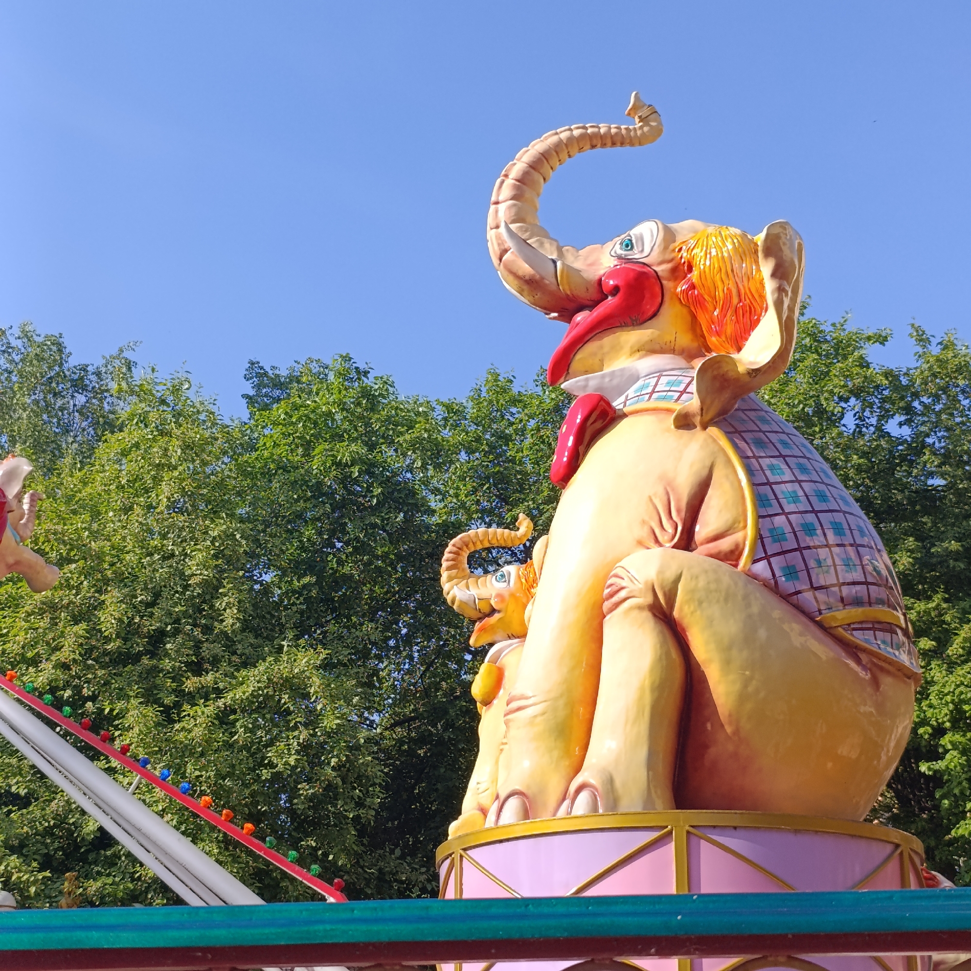 The image shows a colorful elephant sculpture on a carousel ride in Izhevsk, Russia, set against a backdrop of green trees and a clear blue sky. The elephant is primarily yellow, with a red tongue and a checkered pattern on its back. A smaller elephant figure is nestled in front of the larger one. The carousel base is pink and gold. To the left, part of the carousel structure is visible, with colored lights lining a white and red support beam.