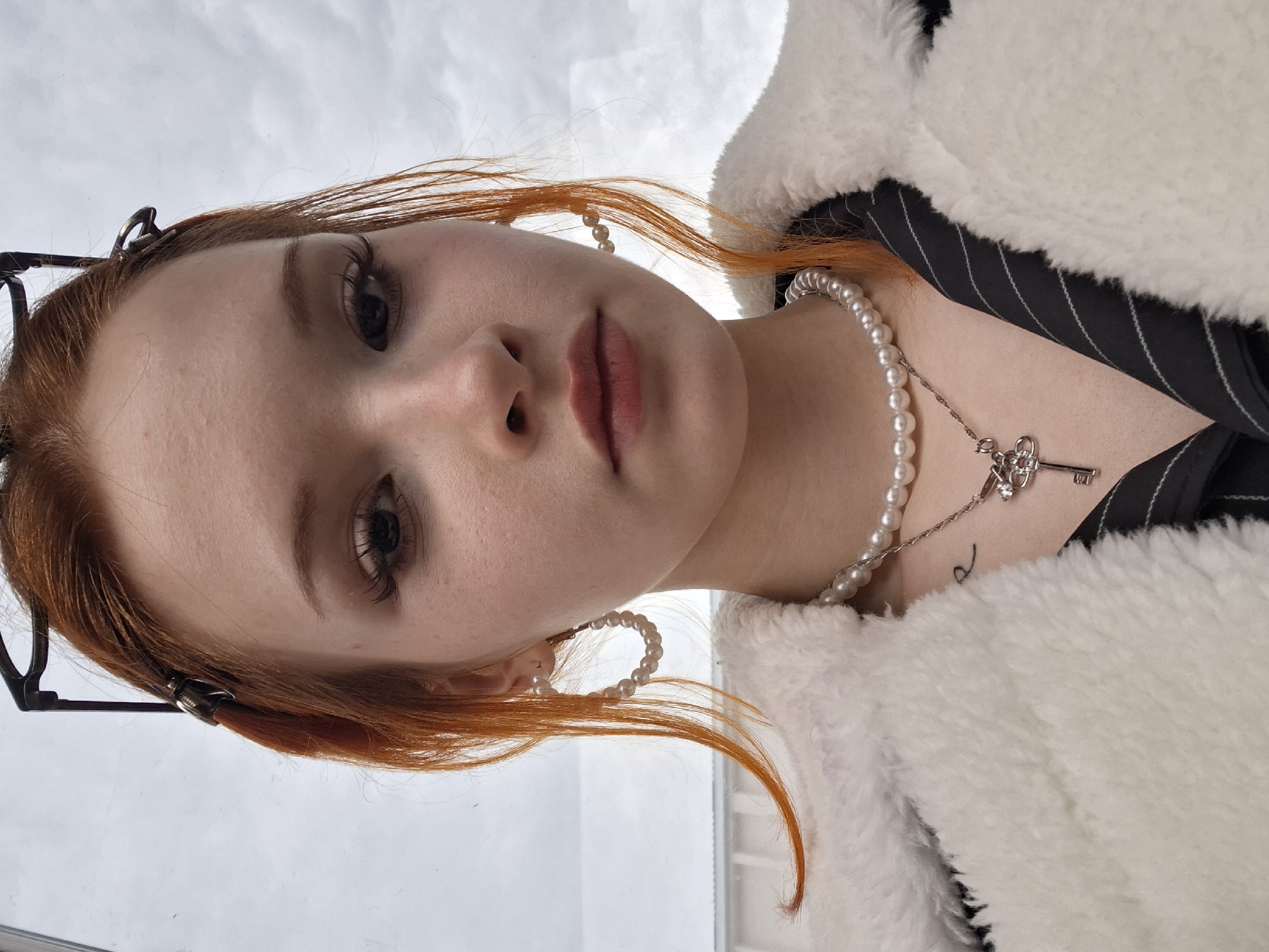 A close-up, tilted selfie shows a fair-skinned person with red hair and dark eyes. They are wearing pearl earrings and a pearl necklace with a key pendant. The person is dressed in a black and white striped garment under a fluffy white coat. The background is indistinct, appearing to be a cloudy sky. There is no explicit evidence indicating this image was taken in Chaikovsky, Russia.