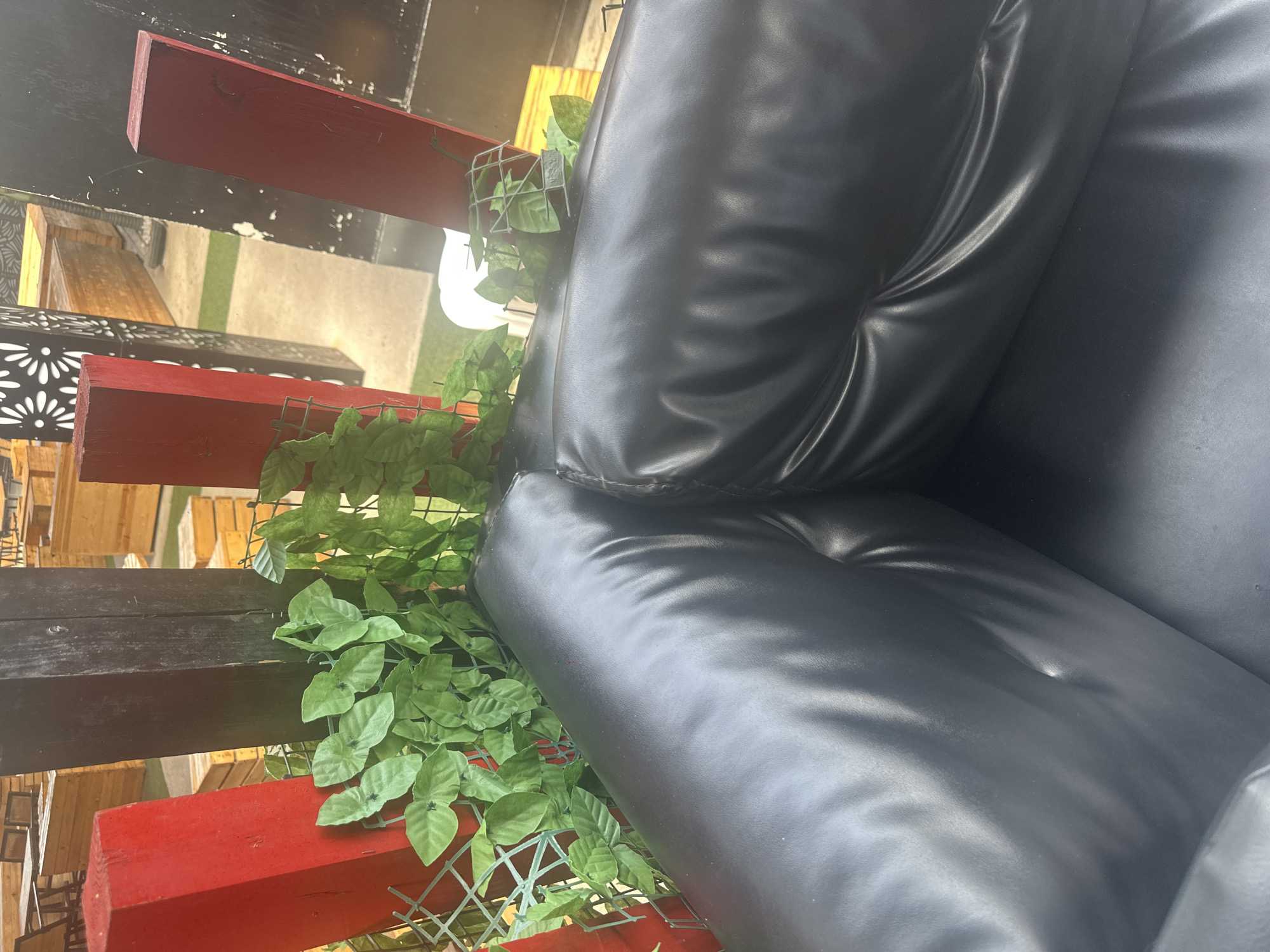 In Howayi, Nigeria, an outdoor seating area features a black leather couch. Behind the couch, several red-painted posts and a dark-colored wooden post are visible, along with artificial green foliage draped over a green wire fence. In the background, wooden benches and tables are arranged on a green turf surface, suggesting a public space like a restaurant or park. The lighting indicates it is daytime.