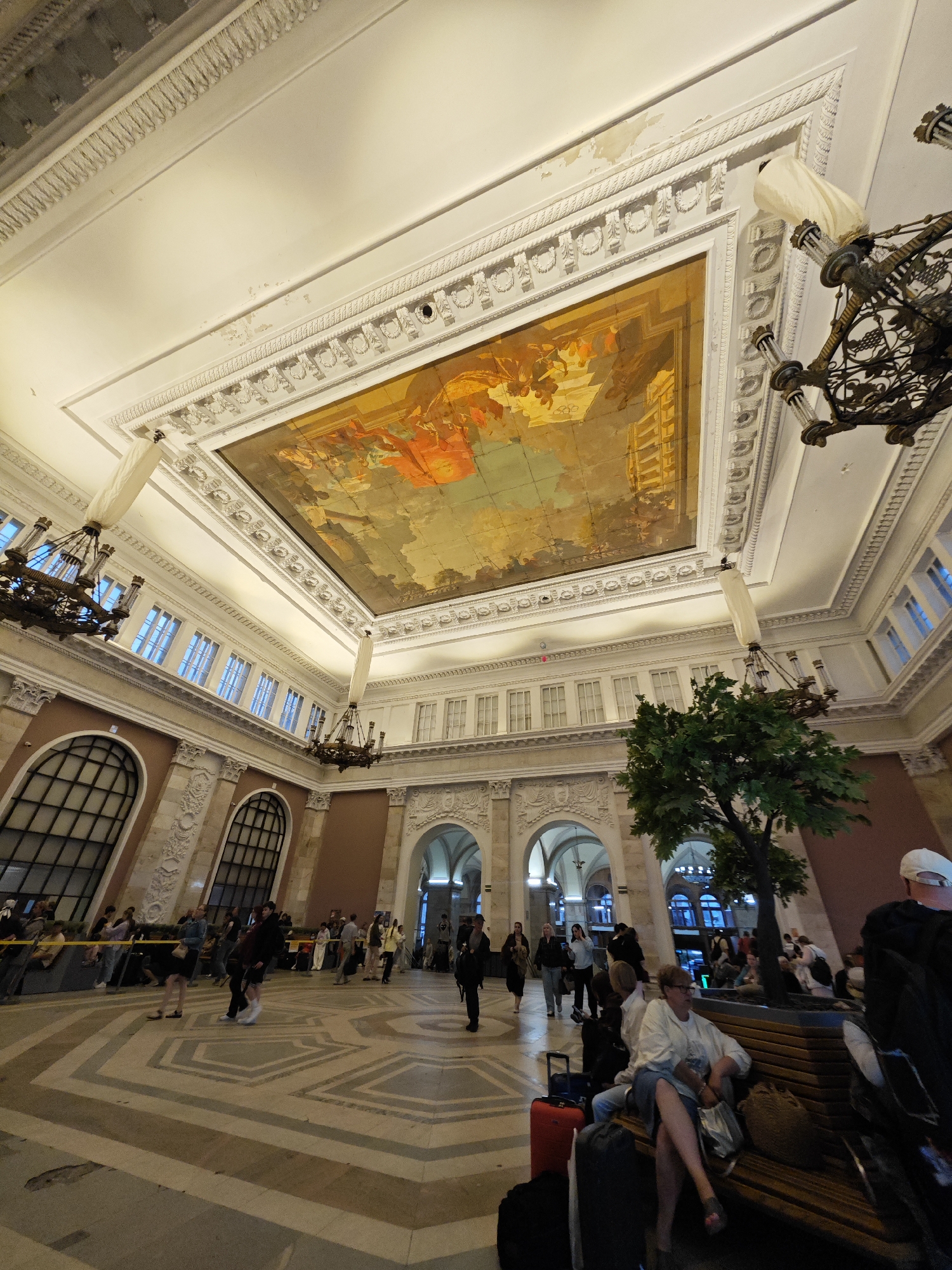 This image captures the interior of the Vitebsky Railway Station in Saint Petersburg, Russia. The station's grand hall is characterized by its high, ornate ceilings, adorned with a large painted mural and elaborate chandeliers. Natural light streams in from large, arched windows along the upper walls. The floor is tiled in a geometric pattern. In the foreground and midground, several people are visible. Some are standing and walking, while others are seated on benches, some with luggage. A faux tree is placed near the center of the hall. The scene appears to be a busy public space, likely during operating hours, with travelers and visitors present. The architecture suggests a historical building, typical of European railway stations.