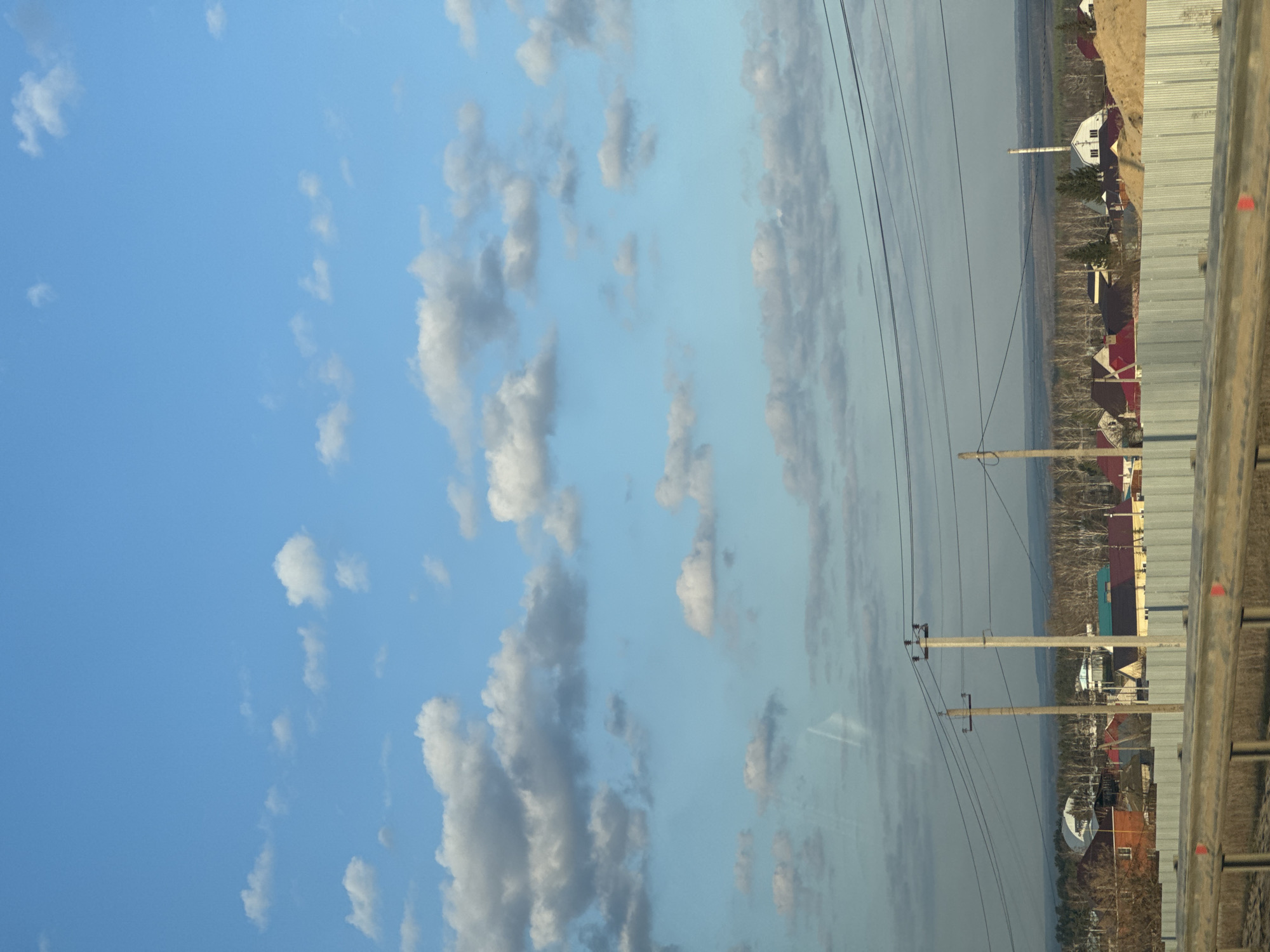 The image shows a view from an elevated position overlooking a town or village. Power lines are visible in the midground. A fence runs along the bottom right of the frame. The sky occupies the left portion of the frame, with scattered clouds. Houses with red and white roofs are visible in the distance. The location is Unknown, Unknown.