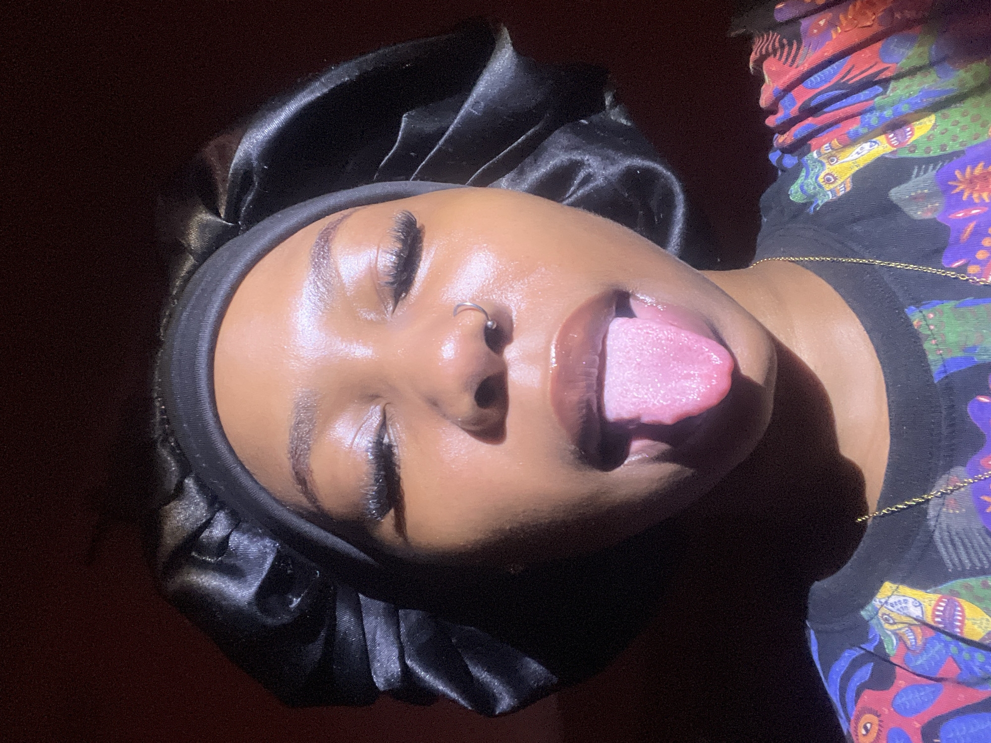 A close-up, selfie-style image shows a young Black woman with her eyes closed and her tongue extended. She is wearing a black satin bonnet and a black t-shirt with a colorful, abstract pattern. A gold chain is visible around her neck. The lighting suggests a single light source from the right, casting a shadow across the left side of her face. The background is a dark, reddish-brown color. There is no visible indication of the location being Rafin Par, Nigeria.