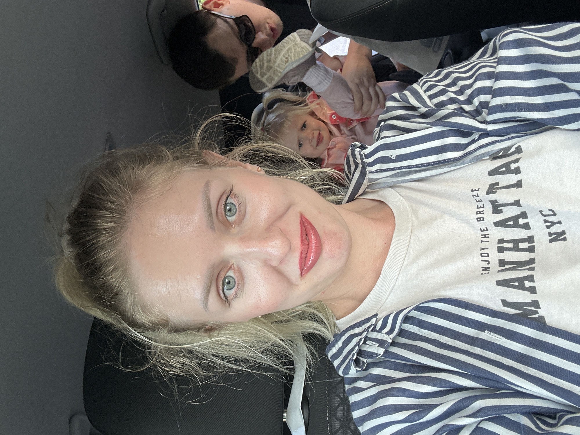 Three people are in what appears to be the back seat of a car. A woman with blonde hair and green eyes is facing the camera. She wears a white t-shirt with "MANHATTAN NYC" printed on it and a striped blue and white shirt. Behind her, a small child with blonde hair and a pink jacket looks towards the camera. Above the child is a man wearing sunglasses. The car's interior is black. The location is not discernible as Petah Tikva from the context of the image.