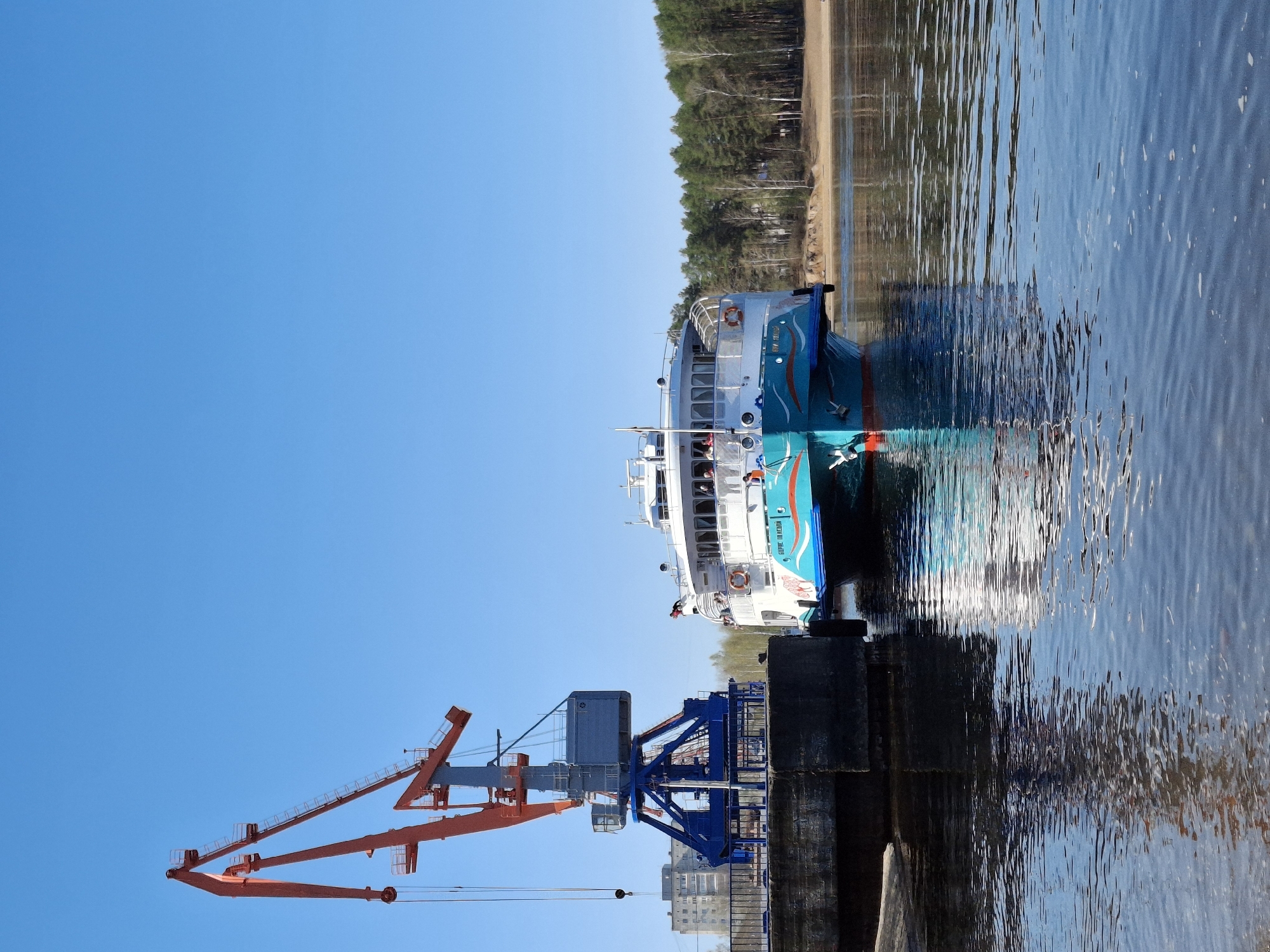 The image depicts a bright blue and white passenger ferry, the "Boris Polevoy," docked at a concrete pier in the city of Chaikovsky, Russia. A large, orange and blue crane stands nearby. The water is rippled, reflecting the sky and the nearby tree-lined shore. A few buildings are visible in the distance beyond the crane.