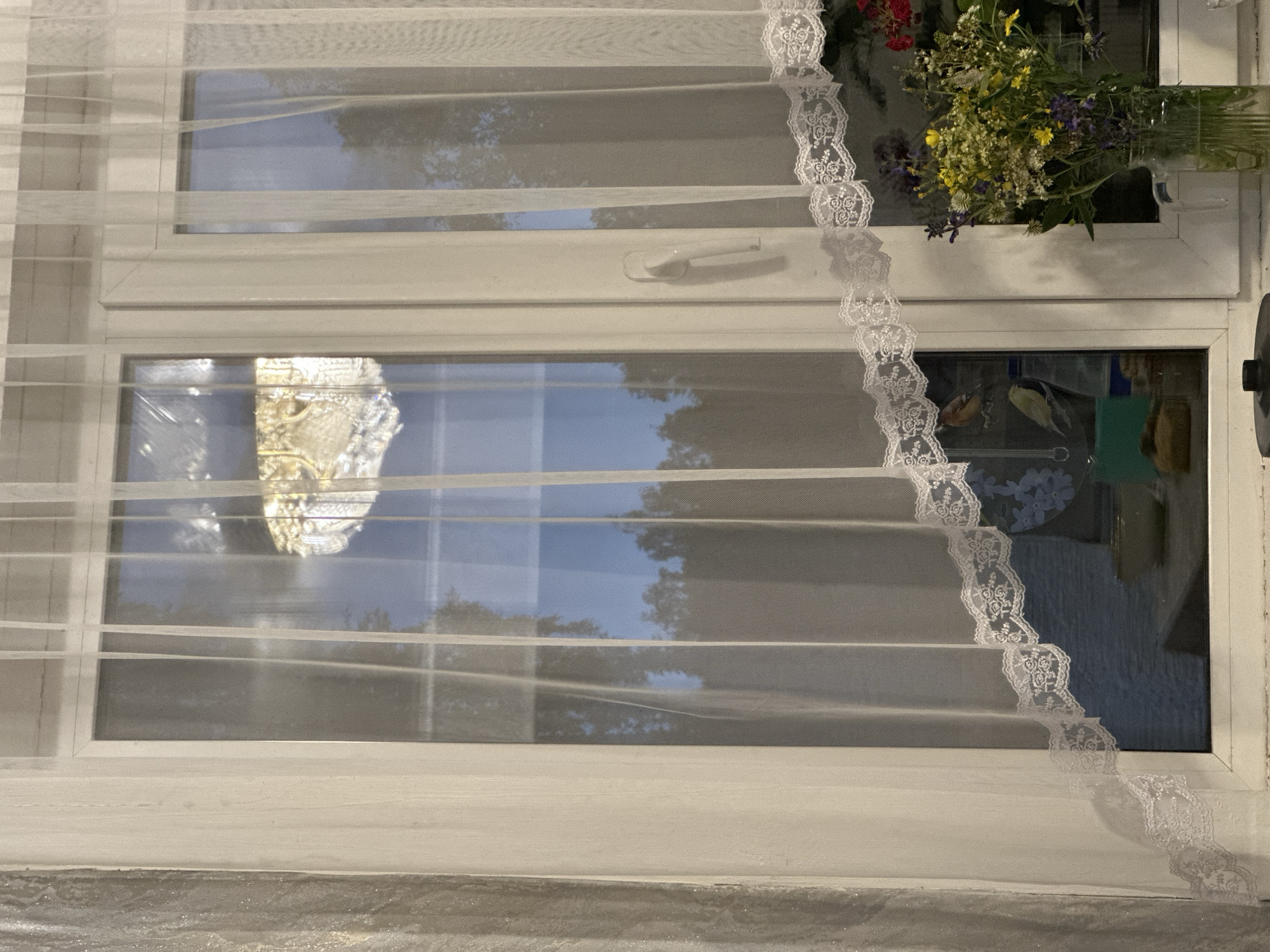 A vertically oriented, medium shot captures a window framed by sheer white curtains. The window itself is divided into two vertical panes, with a white mullion separating them. The left pane reveals a reflection of an ornate chandelier and the twilight sky. The right pane shows a dark silhouette of trees against the sky, with more detailed reflections of the room, including a vase of wildflowers on the right side of the sill. A white window handle is visible on the right pane. The lower portion of the window sill displays various objects, including a green item, a brown item, and a clear glass pitcher filled with a bouquet of wildflowers and greenery. The sheer curtains are adorned with a decorative white lace trim along their bottom edges, which are draped in a wavy pattern across the window. The overall scene suggests an indoor setting looking out through a window, likely during the evening or dusk. The location is Unknown, Unknown.
