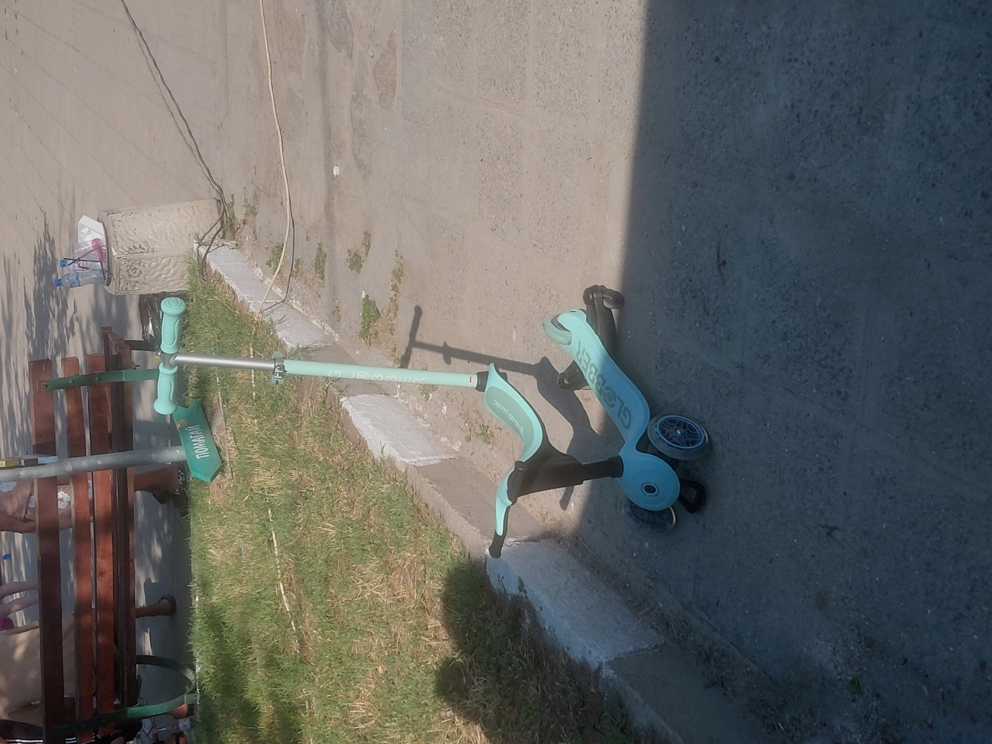 The image depicts an outdoor scene in Kostievo, Bulgaria, under bright daylight. A light blue and black Globber children's scooter, featuring a seat attachment and three wheels, is positioned on a paved surface in the foreground, casting a shadow to its right. To the left, a concrete curb separates the paved area from a narrow strip of dry grass. On this grass, partially obscured, rests a green object, possibly a small board or toy, bearing the Cyrillic inscription "ПОМАГАЙ" (meaning "HELP").
Behind the grassy strip, a wooden park bench with dark metal supports is visible. Several individuals are seated on or near the bench, with only their lower torsos and legs, clad in casual attire and footwear (including sandals), discernable. To the right of the bench, a concrete pedestal with a textured surface functions as a makeshift table, holding two plastic water bottles (one with a blue cap, one with a white cap) and some white cardboard food packaging. Three electrical cords, two dark and one lighter, run across the paved ground, originating near the base of the pedestal and extending out of frame to the right. Strong sunlight from the left casts prominent shadows from the scooter, people, and potential overhead obstructions onto the ground and grass.