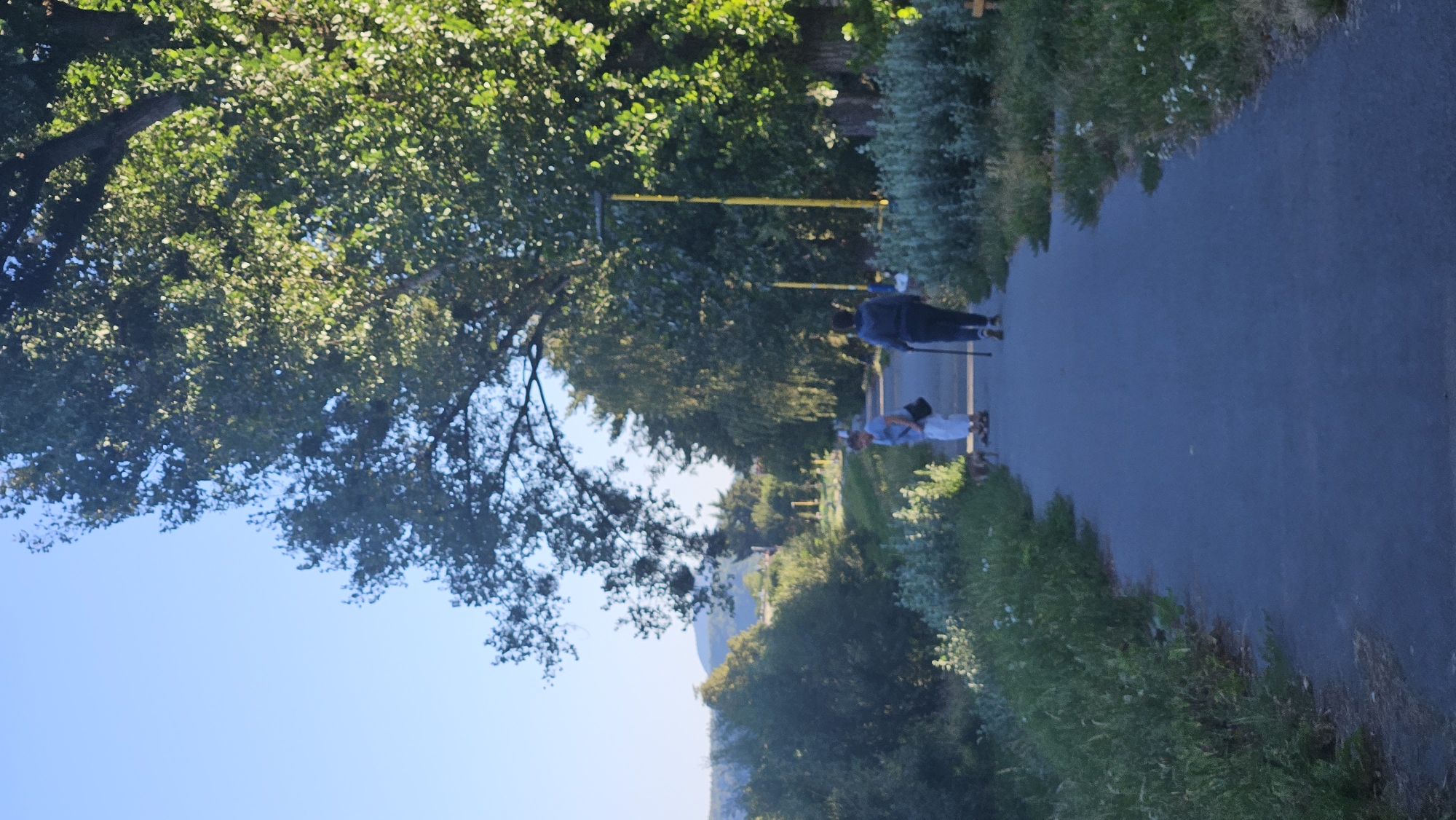The image depicts a paved pathway in a park-like setting, likely in or near Prešov, Slovakia. Two individuals are walking away from the viewer on the path. The person in front, to the right, is wearing a dark blue hooded jacket and dark pants, and is using a walking stick. The person to the left, slightly ahead, is wearing a light blue shirt and white pants, and appears to be with a small dog. The path is bordered by lush green vegetation and trees. In the background, a line of trees forms a backdrop, with distant hills or mountains visible under a clear blue sky. The lighting suggests it is daytime, possibly late afternoon, with sunlight filtering through the leaves of a large tree on the right side of the frame.