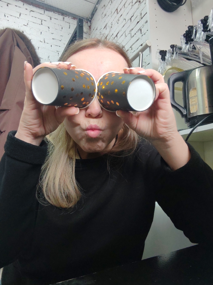 The scene captures an individual, estimated to be a fair-skinned female with blonde hair, directly facing the camera in an indoor setting. She holds two dark gray paper cups, adorned with golden polka dots and white interiors, to her eyes, simulating binoculars. Her mouth is puckered into a pout. She wears a black long-sleeved garment. To the left in the background, a brown coat with a fur-trimmed collar is visible. To the right, a silver and black electric kettle rests on a shelf, above which are clear plastic clips and a black coiled electrical cable. The background wall consists of white-painted brick. The subject is situated before a dark-colored surface, likely a table or counter. The specific location within the city of Chaikovsky, Russia, cannot be determined from the visual information provided within the image.