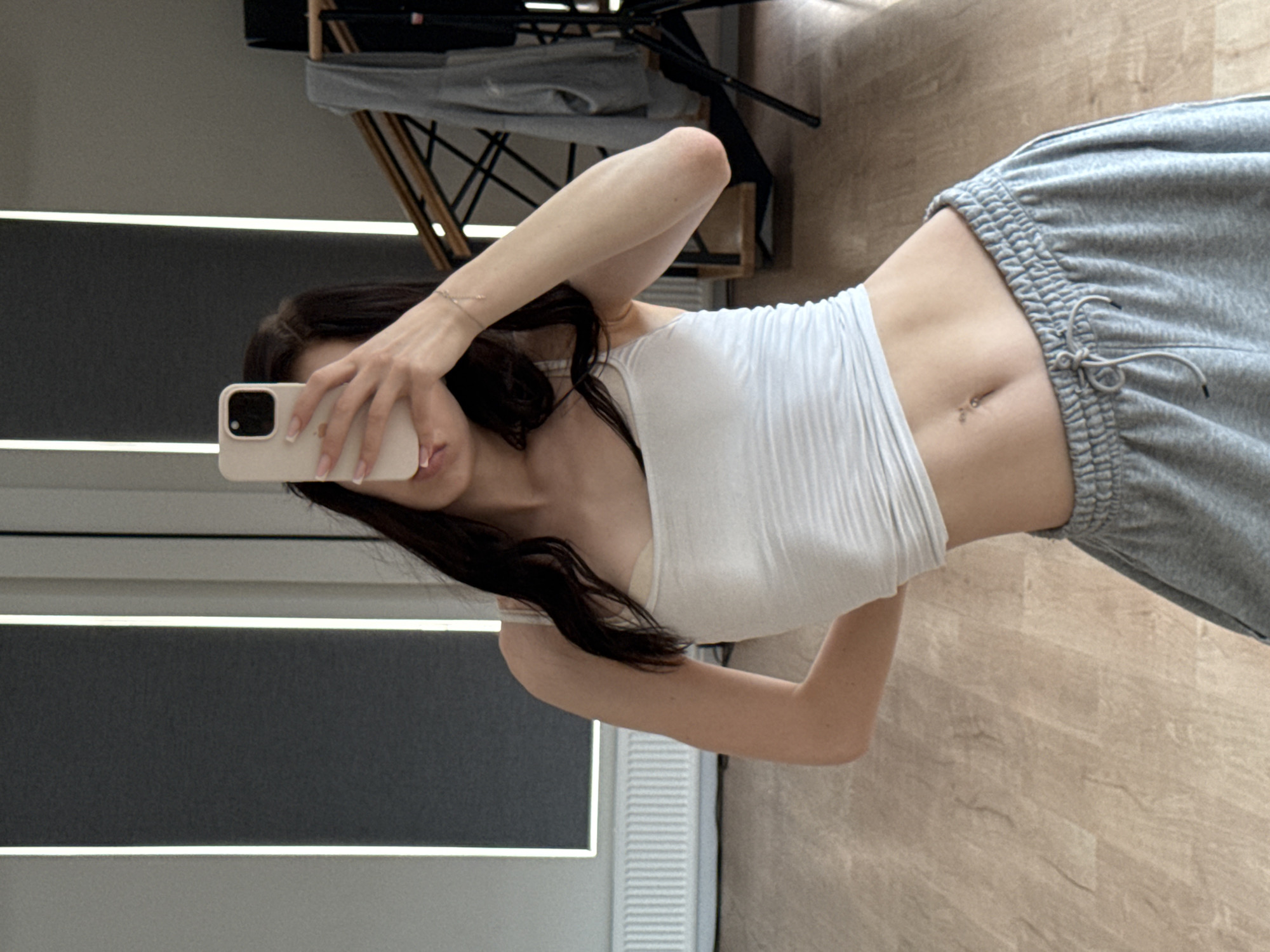 The image shows a young woman taking a mirror selfie in what appears to be a home or studio setting. She is wearing a white tank top and gray sweatpants, with her midriff exposed. She is holding a light-colored smartphone in her right hand, partially obscuring her face. Her left hand is resting on her hip. The background includes vertical blinds, a radiator, wooden flooring, and some furniture, including a wooden structure with a hanging gray garment and a tripod. The environment suggests an indoor space, possibly a room in a residential building or a dance studio. There is no indication of the city of Zakharovo, Russia, or any specific geographical markers within the image.