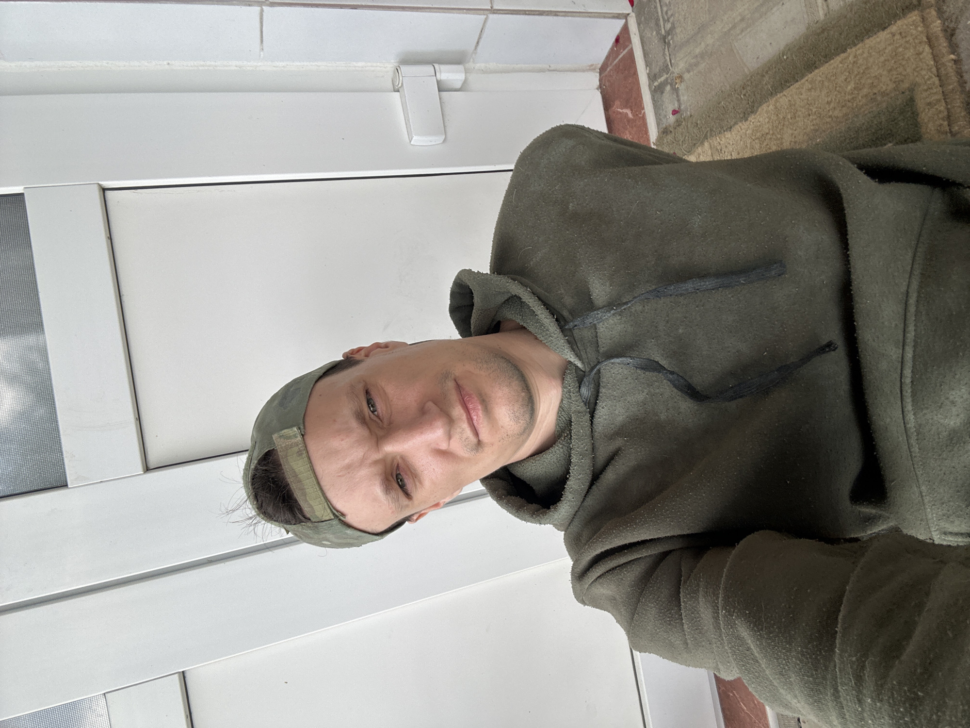 A man wearing a green hooded sweatshirt and a green baseball cap is pictured in a selfie taken outdoors. He appears to be standing or sitting near a white door or window frame. The background consists of white vertical panels and a slightly textured white wall. The man's face is turned towards the camera, and he has a serious expression. The scene appears to be residential.