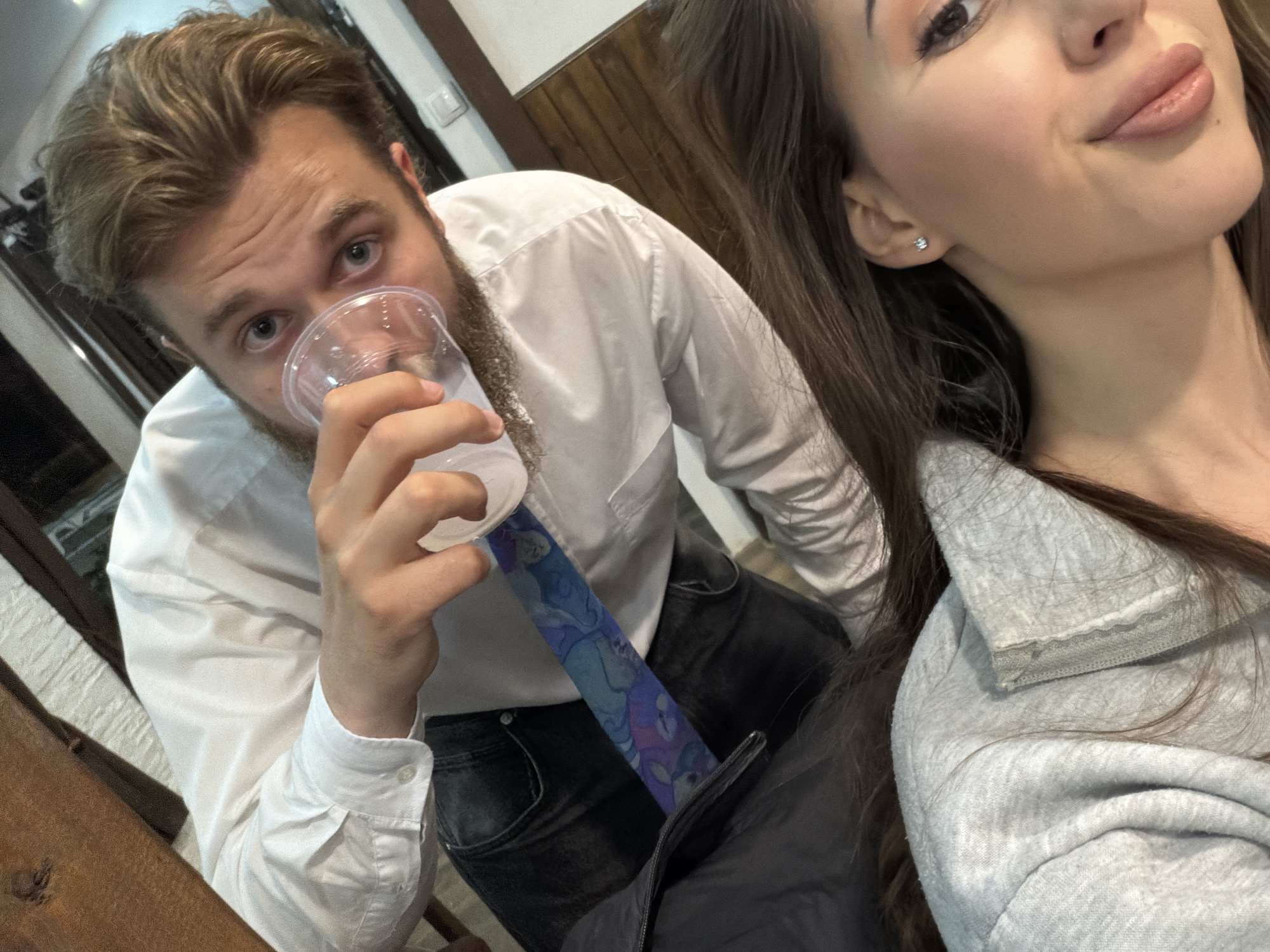 Two people are in the frame. The man on the left is fair-skinned with brown hair and a beard, wearing a white button-down shirt and a blue patterned tie. He is holding a clear plastic cup to his mouth. The woman on the right, also fair-skinned with long brown hair, is wearing a gray sweatshirt.
The background suggests an interior setting with light-colored walls and a wooden panel in the upper right corner.
The location is not discernible as Beli Osam, Bulgaria.