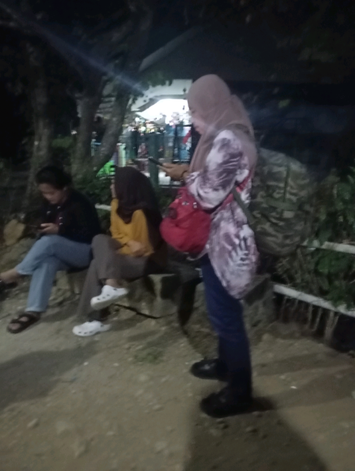 The image depicts a nighttime scene in what appears to be an outdoor public area, potentially a park or a street side in Ketepung, Indonesia. Three individuals are visible, two seated on a bench and one standing.
On the left, a person wearing a black jacket, blue jeans, and dark sandals is seated, looking down at a mobile phone.
In the middle, another person is seated on the bench, wearing a yellow top and dark pants with white Croc-style shoes. This individual is also looking down, seemingly at their phone.
The person on the right is standing and wearing a light-colored hijab and a patterned shirt with a red bag slung over their shoulder. They are also looking down at a mobile phone.
The environment is dimly lit, suggesting nighttime. Trees and foliage are visible in the background, along with what might be a building or structure. The ground appears to be a mixture of dirt and possibly gravel. A low fence or railing is also present in the background. The overall atmosphere is casual, with individuals engaged with their personal devices.