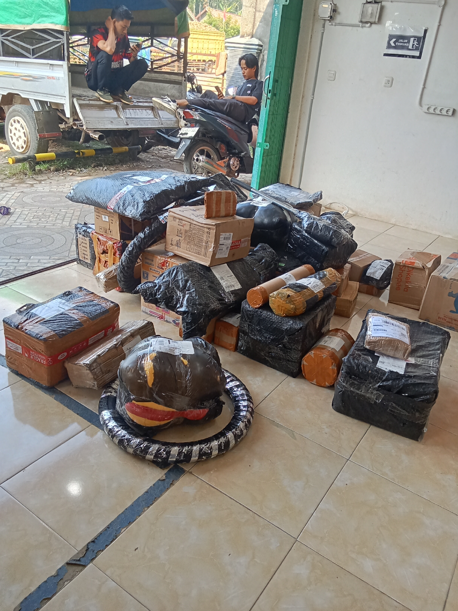 The image depicts a pile of packages and parcels on a tiled floor in front of a building with a white wall. The packages are wrapped in plastic, bubble wrap, and cardboard boxes of various sizes. Some of the packages have shipping labels or markings. In the background, there is a small truck with a green canopy and two individuals, one crouching and looking at a mobile phone, and another sitting on a motorcycle. A sign with "JALUR EVAKUASI" (Evacuation Route) is visible on the wall. The scene appears to be an outdoor area, possibly a loading dock or a delivery point, in Jirak, Indonesia.