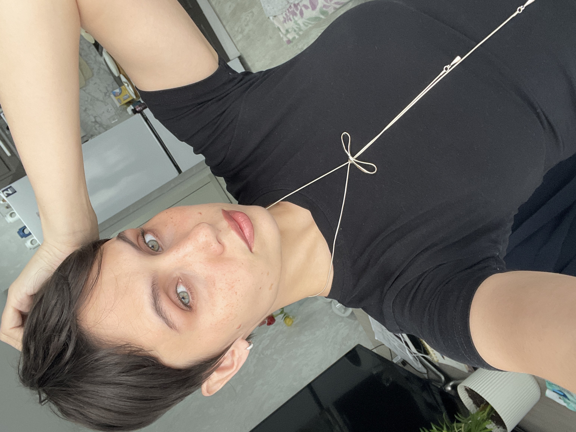 A young adult female is posing for a selfie. She has short, dark hair and light eyes with freckles on her face. She is wearing a black t-shirt. A silver-colored necklace with a small knot is visible around her neck. The background shows a room interior with a white cabinet or refrigerator. A plant in a white pot, a monitor, and other items are also visible. There is insufficient information to identify a specific location in Izhevsk, Russia, or confirm that the image was taken there.