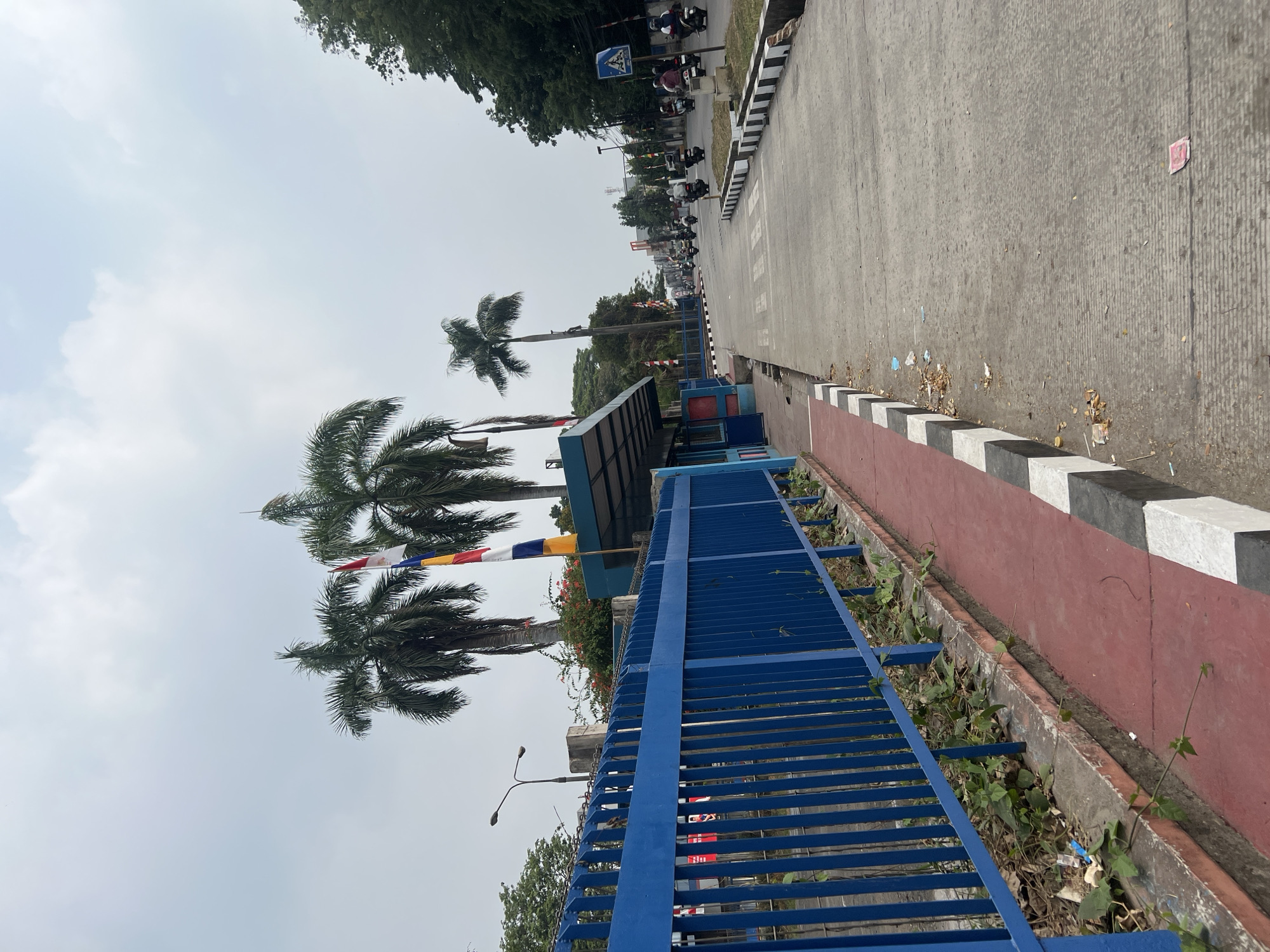 The image shows a street scene in Citeureup, Indonesia, on an overcast day. On the left, a bright blue metal fence runs alongside a red paved walkway. Beyond the fence, several tall palm trees are visible against the sky. A blue building with a covered overhang is situated next to the walkway.
The street itself is paved with asphalt and shows signs of wear and tear, with some debris and fallen leaves scattered on the surface. A pedestrian crossing with black and white markings is present on the road. Several motorcycles, ridden by individuals wearing helmets, are traveling down the street, suggesting moderate traffic. Some trees line the right side of the road, and a pedestrian crossing sign is visible. The overall environment appears to be urban or semi-urban.