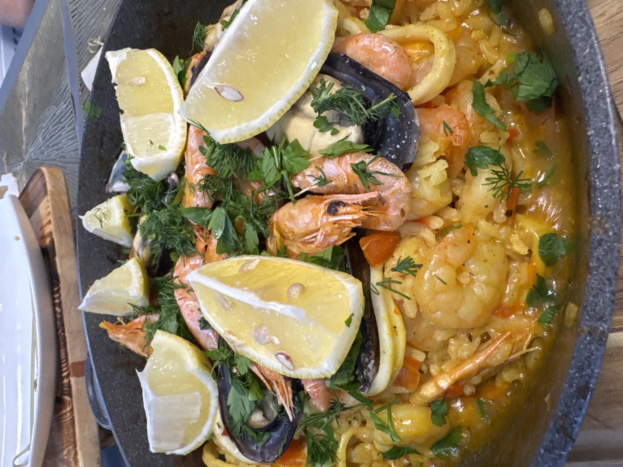 A close-up, overhead view shows a paella dish presented in a dark, wide, shallow pan. The paella is generously filled with rice, interspersed with various seafood including shrimp, mussels, and calamari rings, all coated in a rich, orange-yellow sauce. Bright yellow lemon wedges are strategically placed on top of the dish, along with fresh green herbs, predominantly dill and parsley, which are scattered throughout. The food is served on a wooden surface, with a blurred white plate and a woven placemat visible in the background. The lighting suggests an outdoor or well-lit indoor setting. There are no people visible in the frame. The scene is set in Ладожское Снт, Russia.