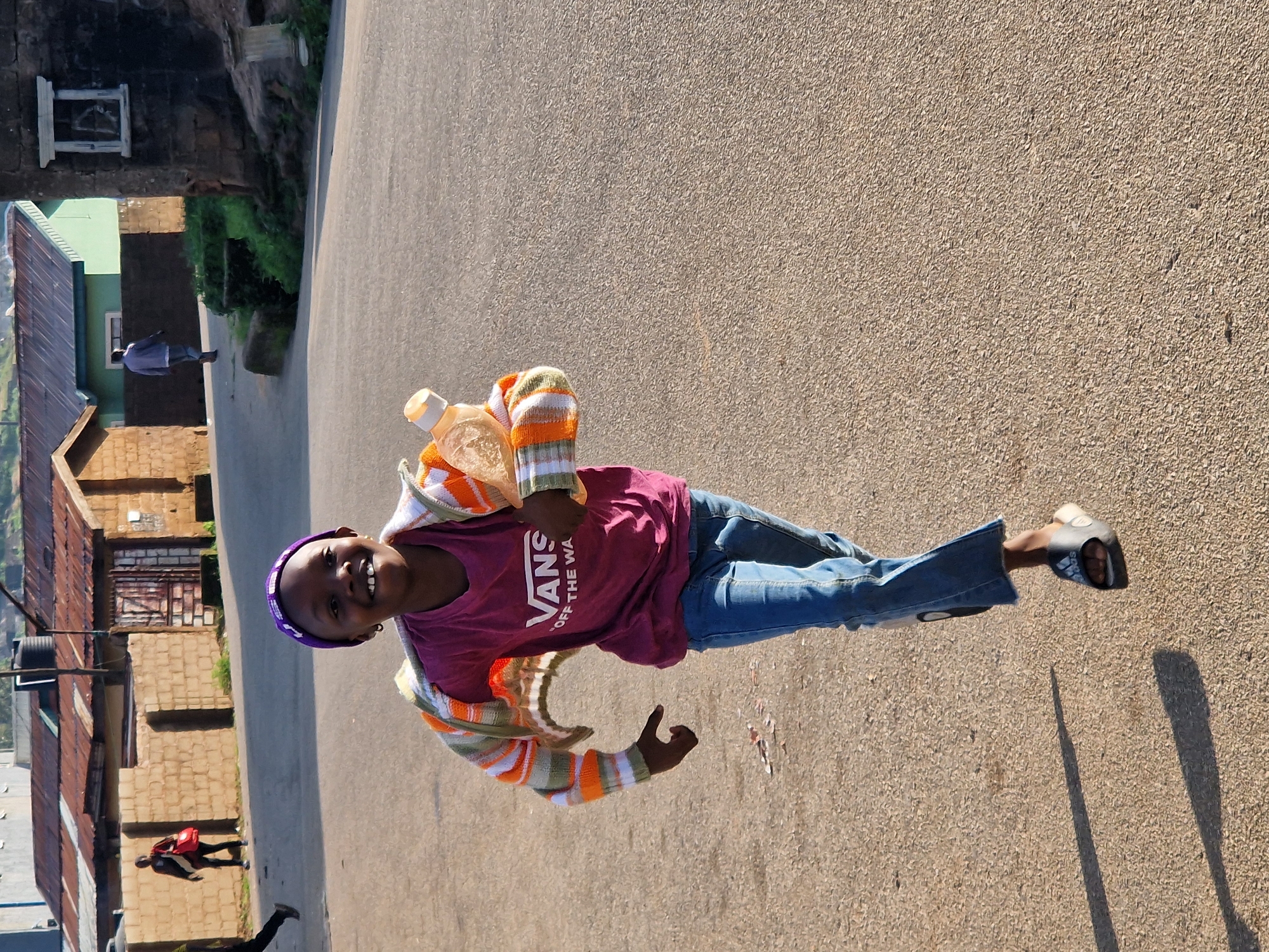 A young girl, wearing a purple beanie, a maroon t-shirt with "VANS OFF THE WALL" printed on it, a striped cardigan, and blue jeans, runs down a paved street in Kabong, Nigeria. She is holding a plastic bottle in her right hand. The girl has a wide smile and is looking towards the camera. In the background, there are buildings with corrugated metal roofs and stone walls, as well as some greenery. Two other individuals are visible in the distance, one walking away from the camera with a red bag, and another walking towards the camera. The scene appears to be lit by natural sunlight, casting shadows on the street.