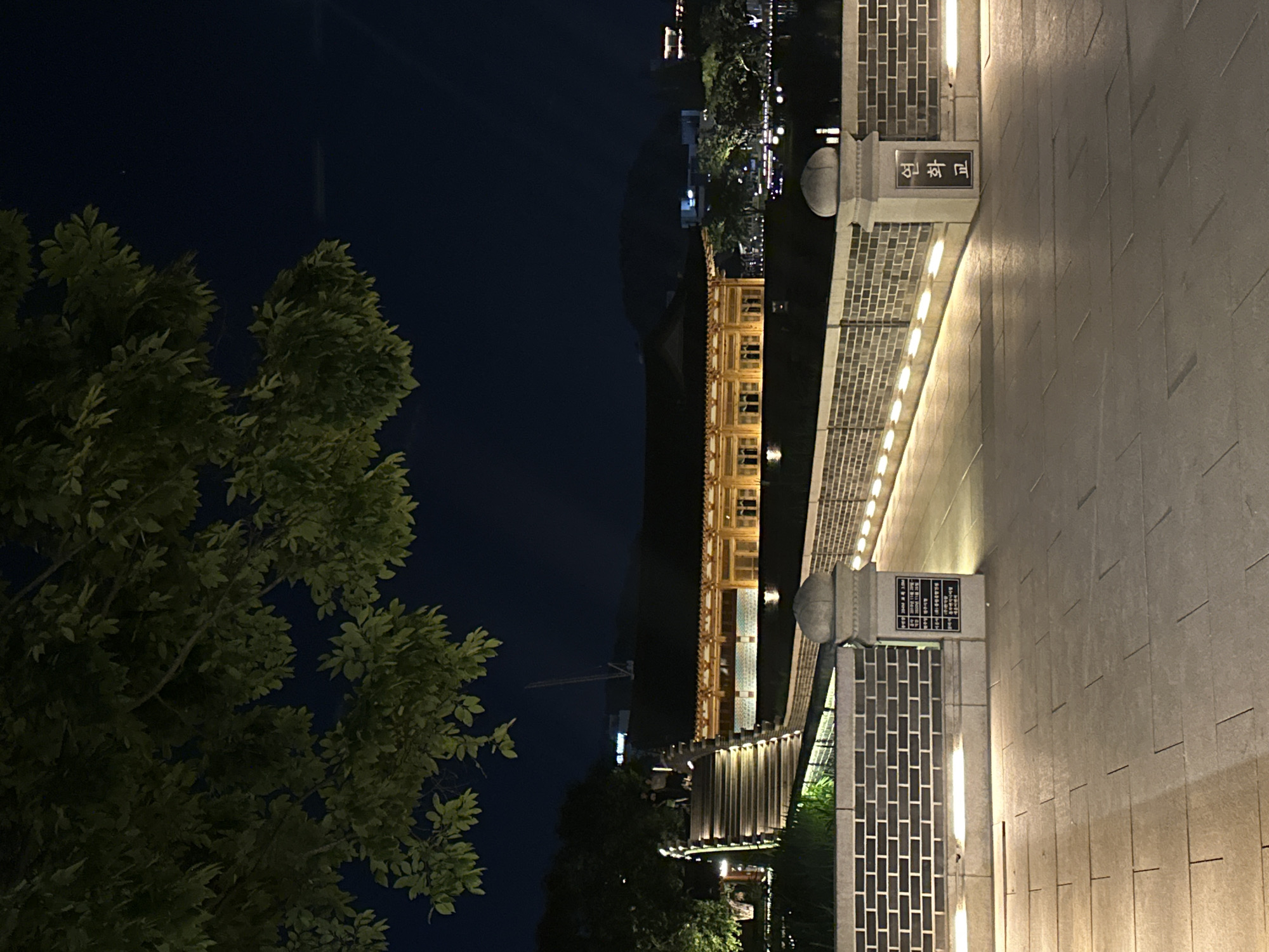 The image depicts an outdoor nighttime scene, likely in the city of Jeonju-si, South Korea, showcasing a traditional Korean architectural structure illuminated by warm lighting. In the foreground, a paved walkway made of large, square stone tiles stretches out, with subtle lines indicating the grout. To the left, a brick wall borders the walkway, with small, upward-facing lights embedded along its base, casting a glow upwards. A stone pillar with a rounded cap stands in front of the brick wall, featuring a sign with Korean text. Further back, a set of wide, illuminated stone stairs ascends to the right, with groups of people visible at the top. Beyond the stairs, a low brick wall continues, also lit from below. A prominent traditional Korean building with a dark, tiled roof and a wooden facade, adorned with multiple windows, is visible in the mid-ground. The building is brightly lit from within and by external lights. In the background, trees with dark foliage are silhouetted against the night sky. Distant city lights and the outlines of hills or mountains are faintly visible. A large tree with green leaves partially obscures the top left portion of the image. The overall atmosphere is serene and illuminated, highlighting the architectural beauty of the location at night.