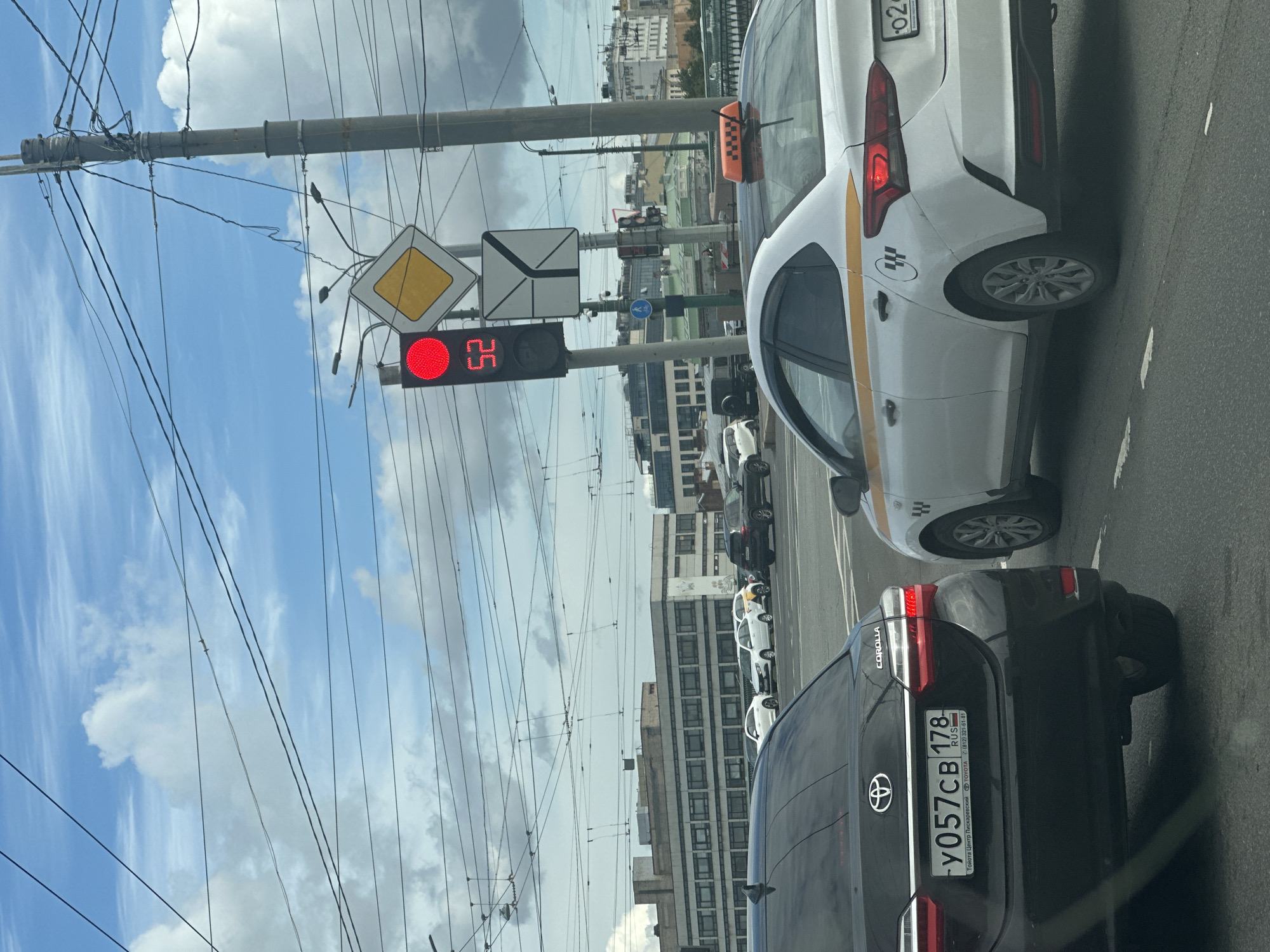 The image shows a view from inside a vehicle looking at a city intersection in Saint Petersburg, Russia. A traffic light is red, and a countdown timer displays "52". Above the traffic light, there is a diamond-shaped yellow sign with a black border, indicating a priority road. Further up, electrical wires crisscross the sky, which is partly cloudy. Several cars are stopped at the intersection, including a black Toyota Corolla with a Russian license plate "y057cb 178" and a white taxi with a yellow stripe and a checkered taxi light on its roof. In the background, buildings line the street, and overhead tram lines are visible. The scene depicts a typical urban traffic situation with elements characteristic of Saint Petersburg, such as the architecture and the presence of trams.