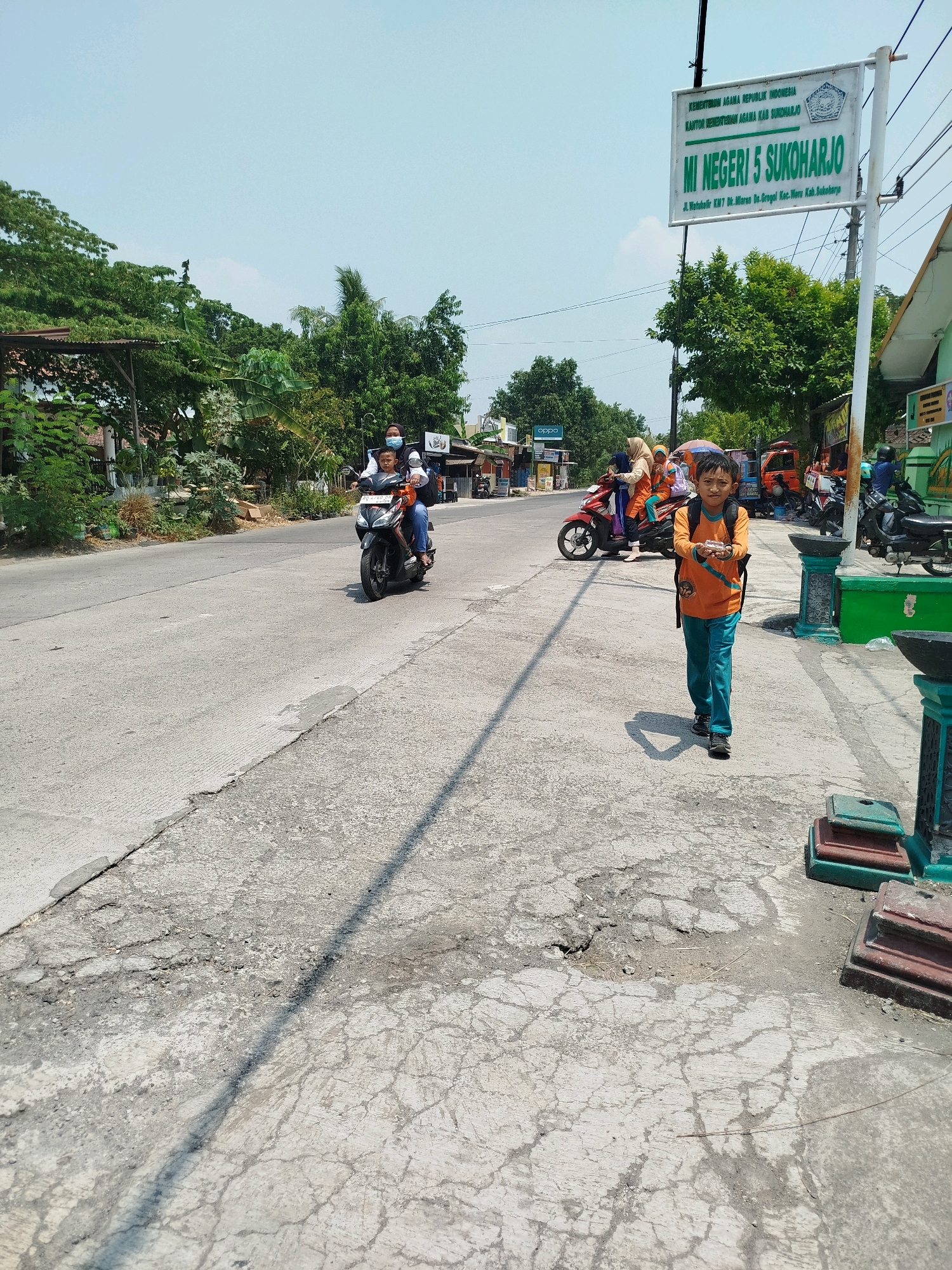 This image depicts a street scene in Grogol, Indonesia, likely outside MI Negeri 5 Sukoharjo. A young boy, wearing an orange long-sleeved shirt and turquoise pants with a backpack, is walking on the sidewalk towards the camera. He is holding a small, light-colored object in his right hand. Behind him, a woman is riding a motorcycle with a child in front, and several other people and motorcycles are visible in the background. A sign for "MI NEGERI 5 SUKOHARJO" is prominent on the right side of the image. The street appears to be paved with concrete and shows signs of wear and tear, with cracks and patches visible. Trees and buildings line the street, suggesting a suburban or semi-urban environment. The sky is clear and bright, indicating a sunny day.