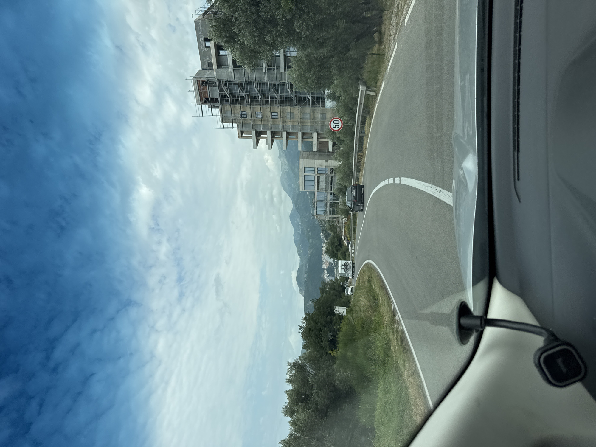 The image, taken from a vehicle's interior, depicts a scenic road curving to the right in Podličak, Montenegro, during daylight hours under a partly cloudy sky. The asphalt road features clear white lane markings and a solid white line delineating its edge.
On the left side of the road, a large multi-story building is under active construction, heavily covered with metal scaffolding. The concrete structure shows multiple window openings. Behind this building, other structures and a range of mountains are visible in the distance. A red and white circular speed limit sign, displaying "50," stands prominently by the roadside near the construction site. Further ahead on the road, a dark-colored car, possibly an SUV, can be seen.
The foreground on the right reveals part of the interior of the car from which the photo was taken, including a section of the dashboard and a black charging cable or phone mount attached within the vehicle. Green vegetation, comprising trees and grass, lines the roadside, adding to the natural landscape. The sky transitions from a deep blue on the far left to a lighter, cloud-dappled expanse above the mountains.
The only clearly legible text is the number "50" on the speed limit sign. The overall scene suggests a journey through a developing coastal or mountainous region.