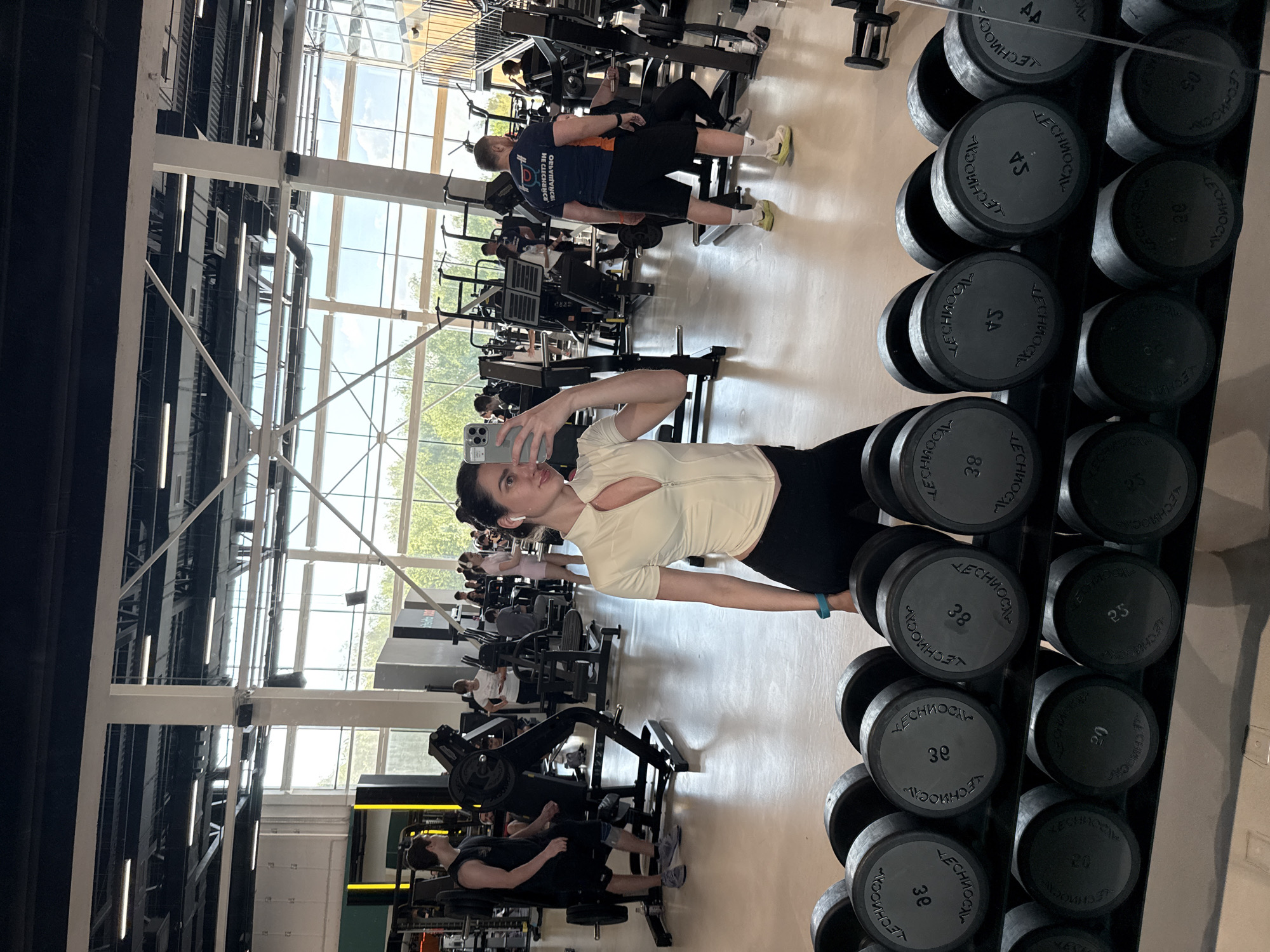 Here is a forensic description of the image:
**Scene:** The image was taken in a gym. It is well-lit with natural light from large windows visible in the background.
**People:** A woman is taking a selfie in the foreground. She is wearing workout clothes consisting of a white top and black leggings. In the background, there are multiple people using different workout machines and equipment, mostly men.
**Objects:** A rack of dumbbells is prominently displayed on the right side of the image. The weights vary in size and have labels on them. Various gym machines are visible in the background, including leg presses. A smartphone is held up by the woman in the foreground.
**Actions:** The woman is holding her phone and taking a picture of herself. The other people in the gym are engaged in various workout activities using the equipment.
**Interactions:** The woman appears to be the central focus of the image, while the other individuals are engaged in their own activities in the background.
**Environment:** The environment seems to be a public gym space, with multiple users and equipment for various exercises.