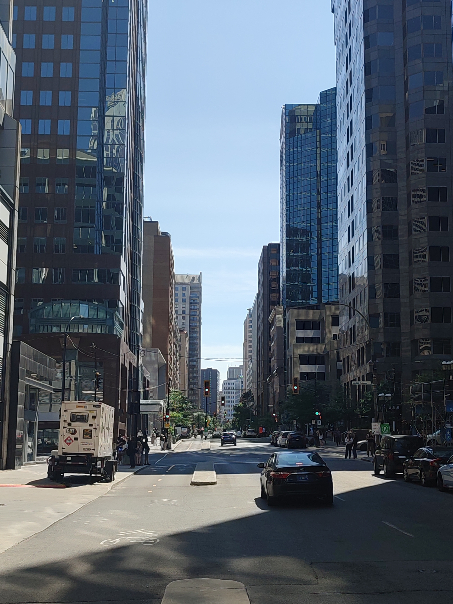This is a street-level, eye-level, vertical, full-shot photograph of a city street in Montreal, Canada, on a clear, sunny day. The street is lined with numerous tall, modern office buildings with reflective glass facades. The buildings on the left are a mix of dark brown and lighter grey tones, while those on the right are predominantly dark grey. In the foreground, the street is paved and appears to have a designated bicycle lane marked with a white symbol.
On the left side of the street, a white trailer with a "CAT" logo is parked. Several people, dressed in casual attire, are standing and walking on the sidewalk near the trailer. Traffic lights are visible at intersections, with some showing green and others red.
In the middle ground and background, cars are driving on the street. A black sedan is prominent in the right lane of the foreground. Further down the street, other vehicles are visible, as are more pedestrians on the sidewalks. Trees and streetlights are interspersed along the street. The sky is a clear, light blue. The scene depicts a typical urban environment with commercial activity and pedestrian and vehicular traffic.