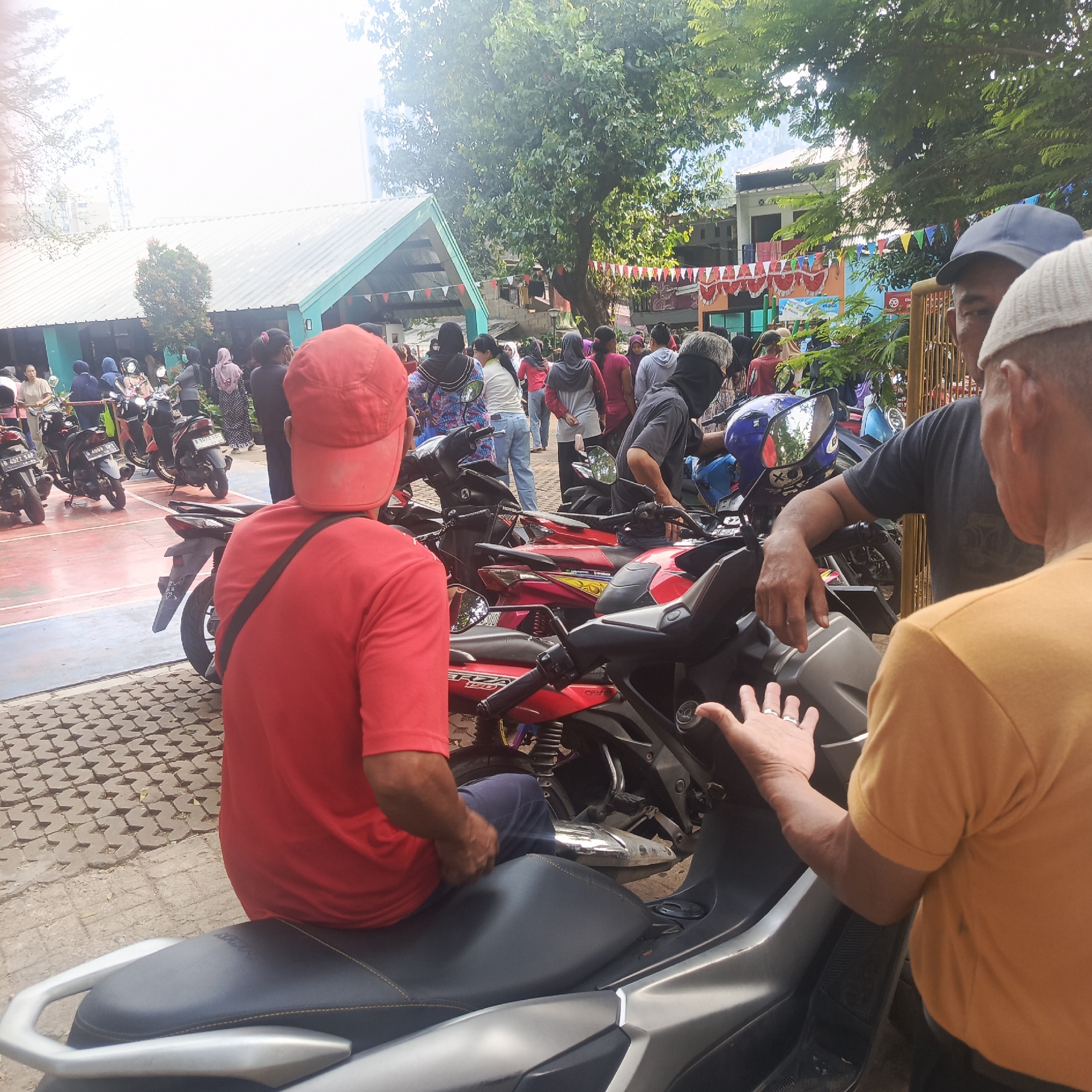 The image depicts an outdoor scene likely in South Jakarta, Indonesia, possibly on a paved court or open area adjacent to buildings. A group of people, predominantly dressed in casual attire and some wearing headscarves, are gathered around a collection of parked motorcycles. The atmosphere appears to be lively, with a sense of community or an event taking place. Red and white triangular flags are strung across some of the buildings, suggesting a celebratory occasion, possibly Independence Day.
In the foreground, a person wearing a red t-shirt and a red cap is seated on a motorcycle, viewed from the back. To their right, another person, seen from the side and back, is wearing a dark gray t-shirt and a black face mask, and is interacting with the person in the foreground. A third individual, wearing an orange t-shirt and a white cap, is partially visible on the right side of the image, also engaged in conversation or interaction.
The motorcycles are of various models, with some parked in a row, and others clustered together. The ground is a mix of paved interlocking blocks and a lighter colored surface, possibly a sports court. In the background, there are low-rise buildings with distinctive green and teal trim on their roofs, and mature trees providing shade. The overall lighting suggests it is daytime, with bright sunlight. The presence of multiple motorcycles and the gathering of people could indicate a local community event, a parking area for a venue, or a motor vehicle-related gathering.