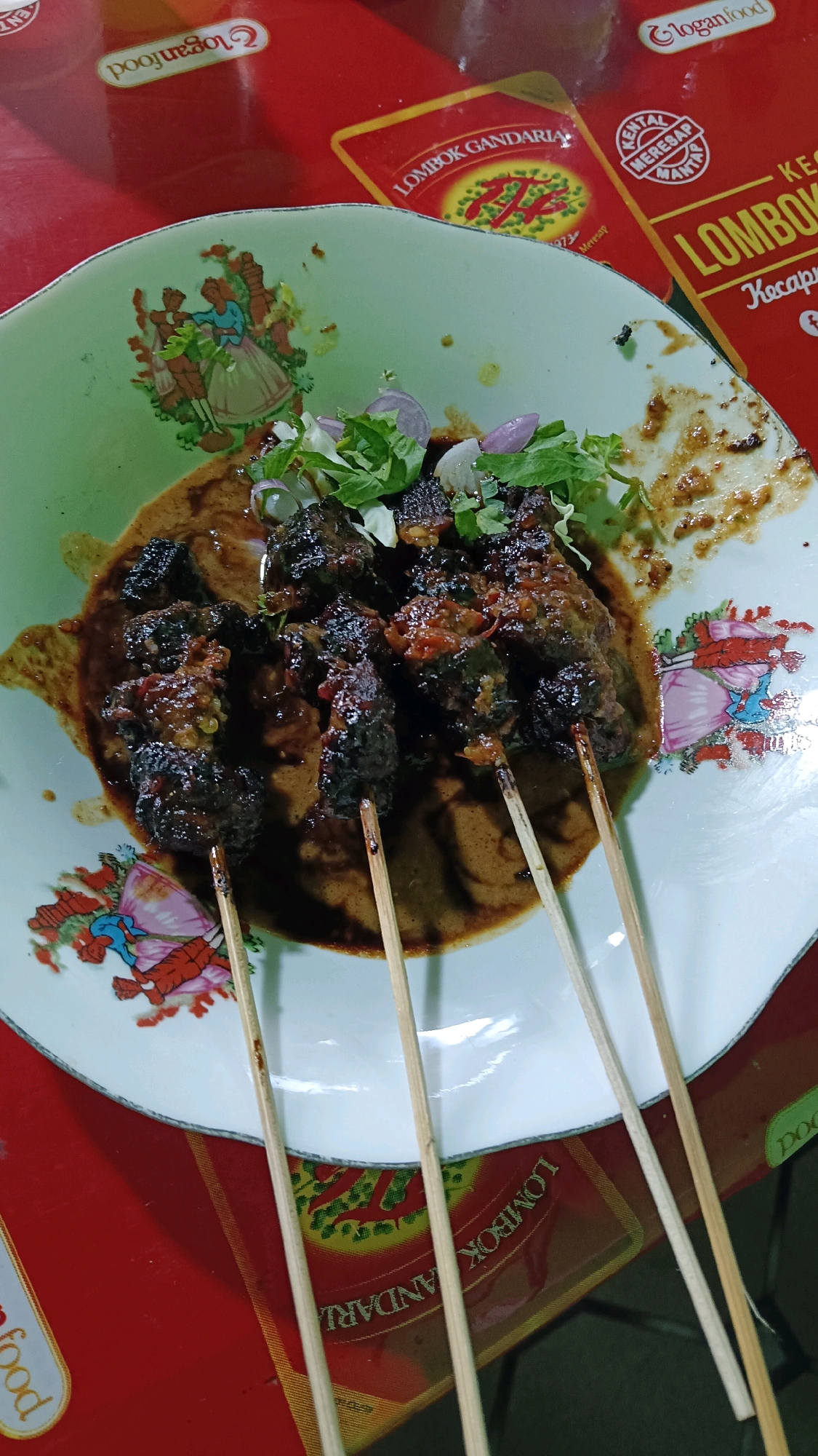 A close-up, top-down view captures a plate of meat satay with a rich, brown sauce and a garnish of chopped onions and green herbs, resting on a red tablecloth. The plate, adorned with a delicate floral pattern featuring figures in traditional attire, sits on a red surface. Behind the plate, two rectangular food packaging labels are visible, one featuring the text "Lombok Gandaria" and the other "Lombok Kecap". The packaging suggests a location in Indonesia, possibly related to the food product. The scene is illuminated by ambient light.