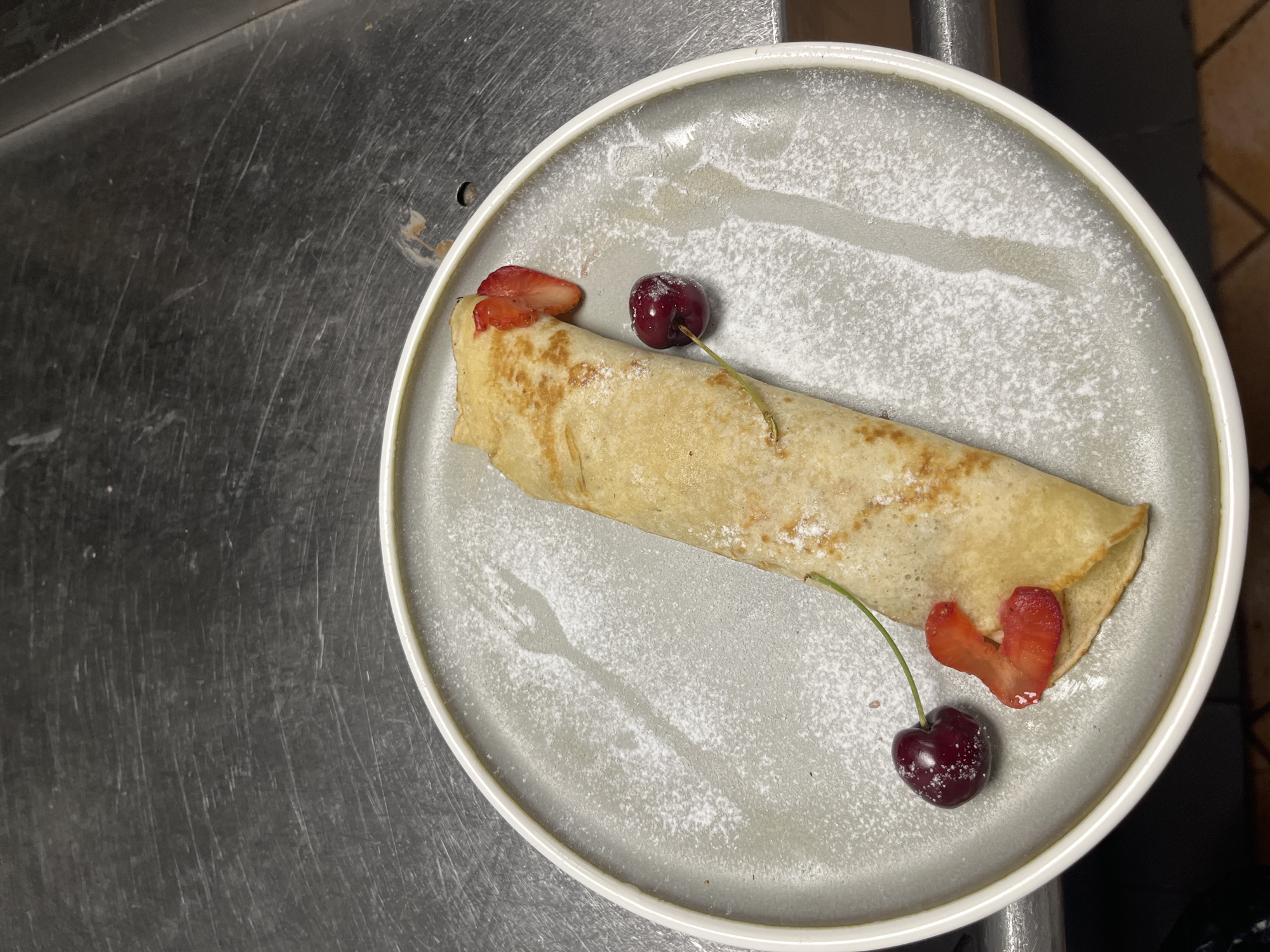 A close-up, overhead shot captures a single, rolled crepe on a gray, speckled plate. The crepe is dusted with powdered sugar, creating a delicate white coating. Two dark red cherries and two heart-shaped slices of strawberry adorn the crepe, adding pops of color. One cherry rests near the top-left of the crepe, with a small piece of strawberry beside it. Another cherry sits near the bottom-right, with a similar strawberry slice. Both cherries still have their thin green stems attached. Powdered sugar is also sprinkled in a light dusting around the plate, with a subtle impression of a fork visible on the left side.
The plate itself has a slightly raised white rim and is placed on a dark, metallic surface that appears to be a countertop. The surface is textured with numerous fine scratches, suggesting it's a well-used workspace. The lighting appears to be artificial, casting a soft glow on the food and plate. The overall scene is a simple and elegant presentation of a dessert. No people are visible in the frame. The time of day and weather are not discernible from the image. There is no visible text in the image.