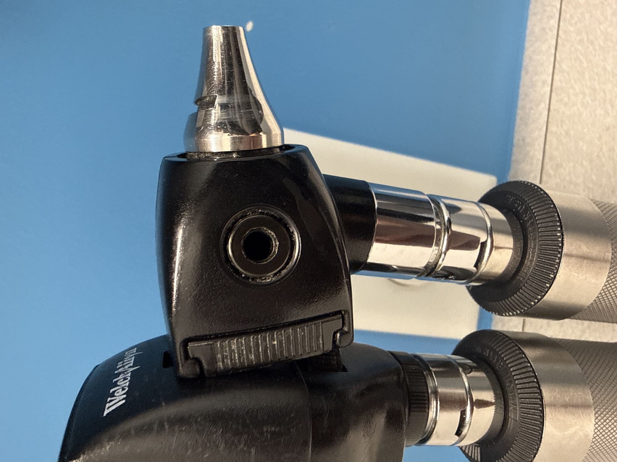 The image shows a close-up of two Welch Allyn otoscopes. The otoscopes are black and silver in color. The top otoscope has a silver speculum attached. The bottom otoscope has the Welch Allyn logo visible. The background is a light blue wall. The location is likely a medical office in Hilliard, United States of America.