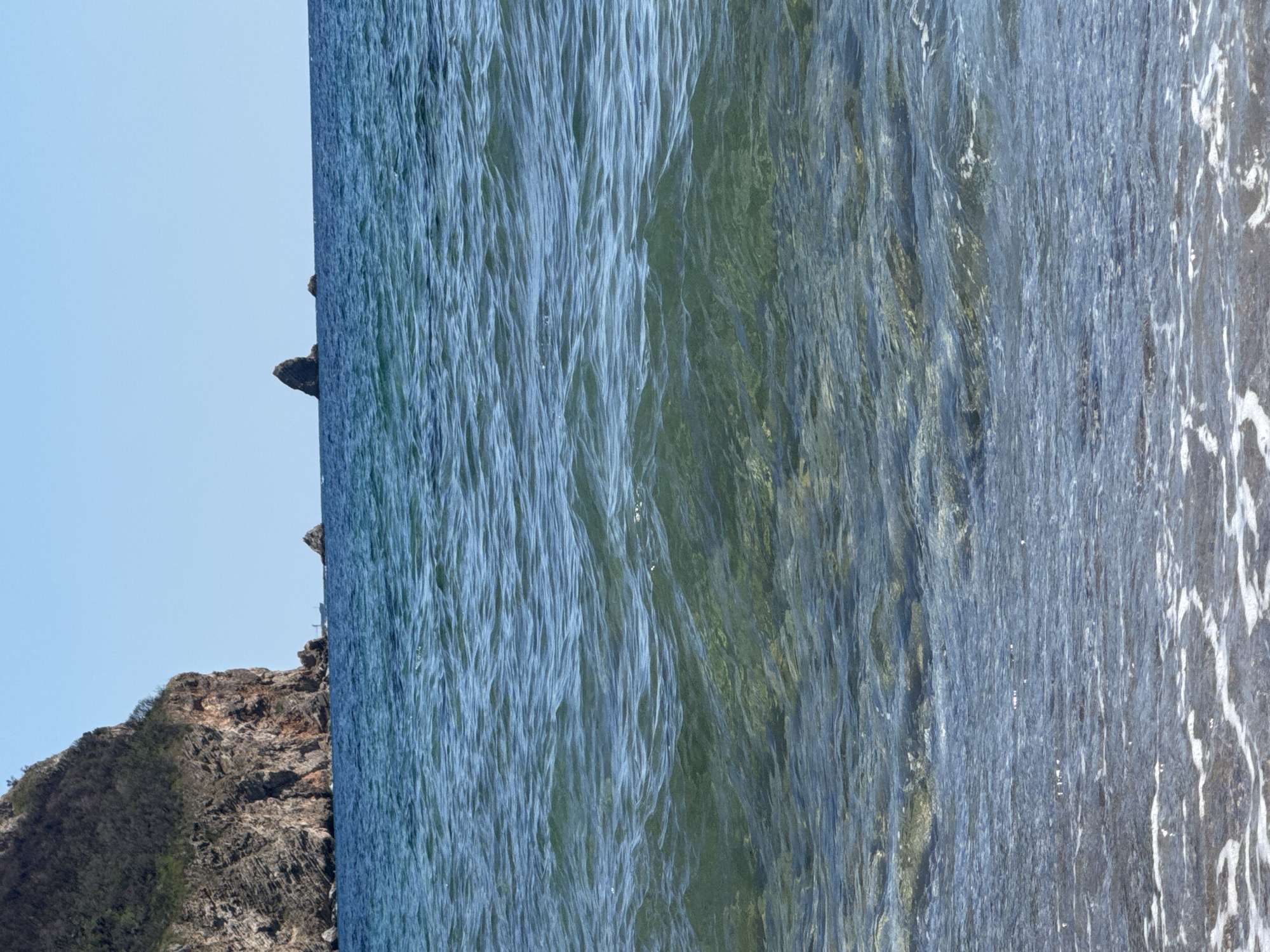 The image depicts a body of water with a rocky cliff on the left and a clear blue sky. In the distance, there are two rock formations emerging from the sea. The water is a gradient of blues and greens, with visible ripples and some white foam at the shore in the foreground, suggesting a beach. The cliff on the left is steep and covered with vegetation on its upper slopes. A small boat is visible near the base of the cliff. The overall scene suggests a coastal environment in Đurovići, Montenegro, likely a scenic overlook or beach area. There are no people visible in the image.